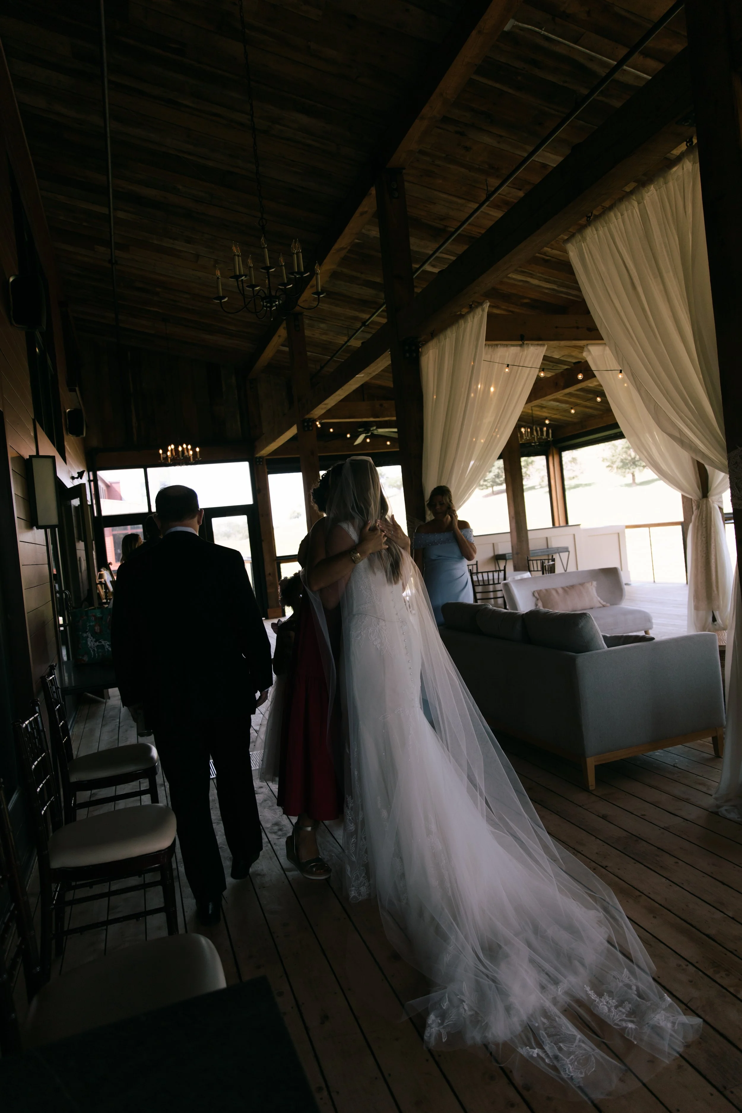 A bride embracing a guest in a rustic indoor venue with wooden floors, curtains, and soft lighting.