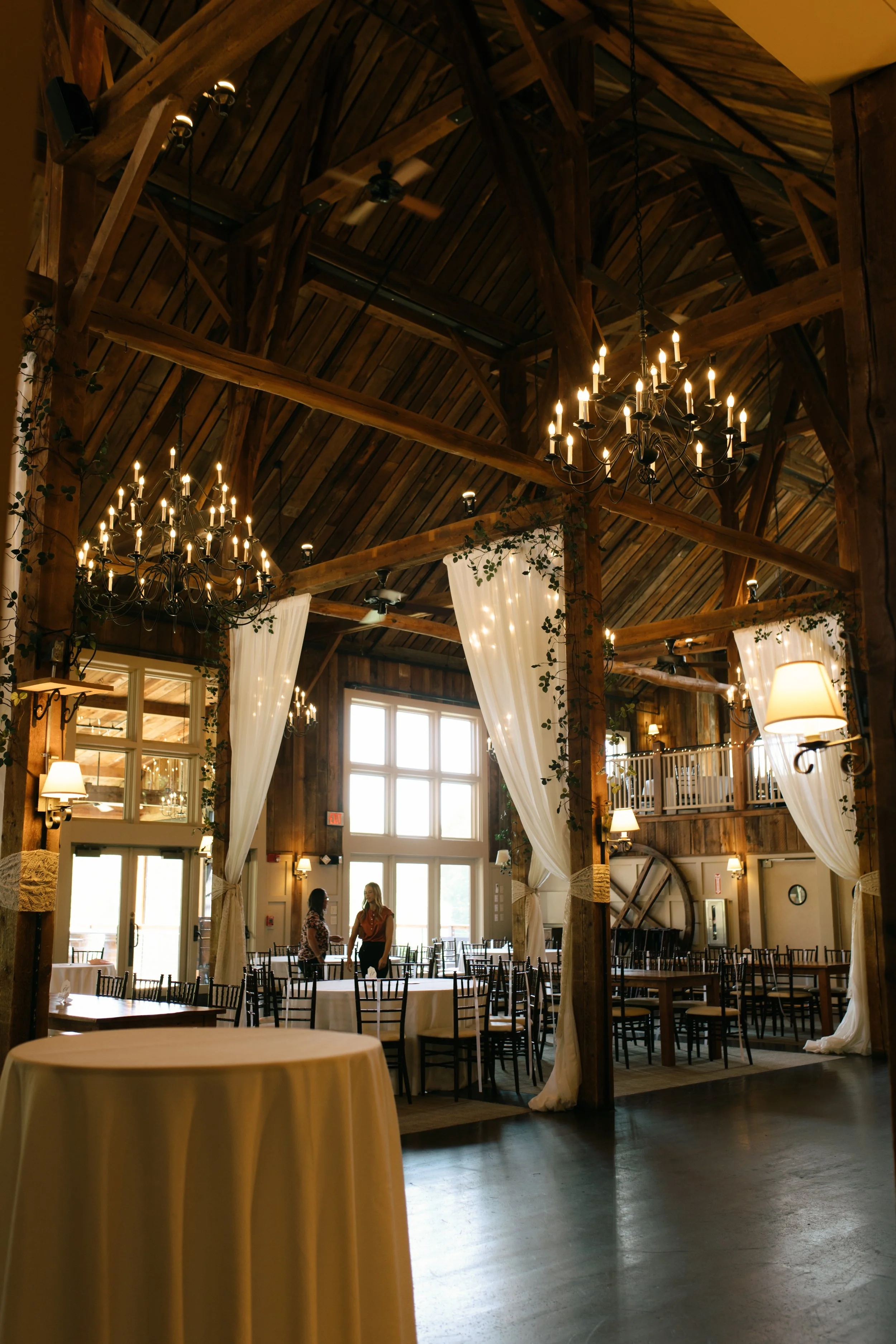 Interior of a rustic event venue with high wooden ceilings, chandeliers, white curtains, and decorated tables, with two women talking near a large window.