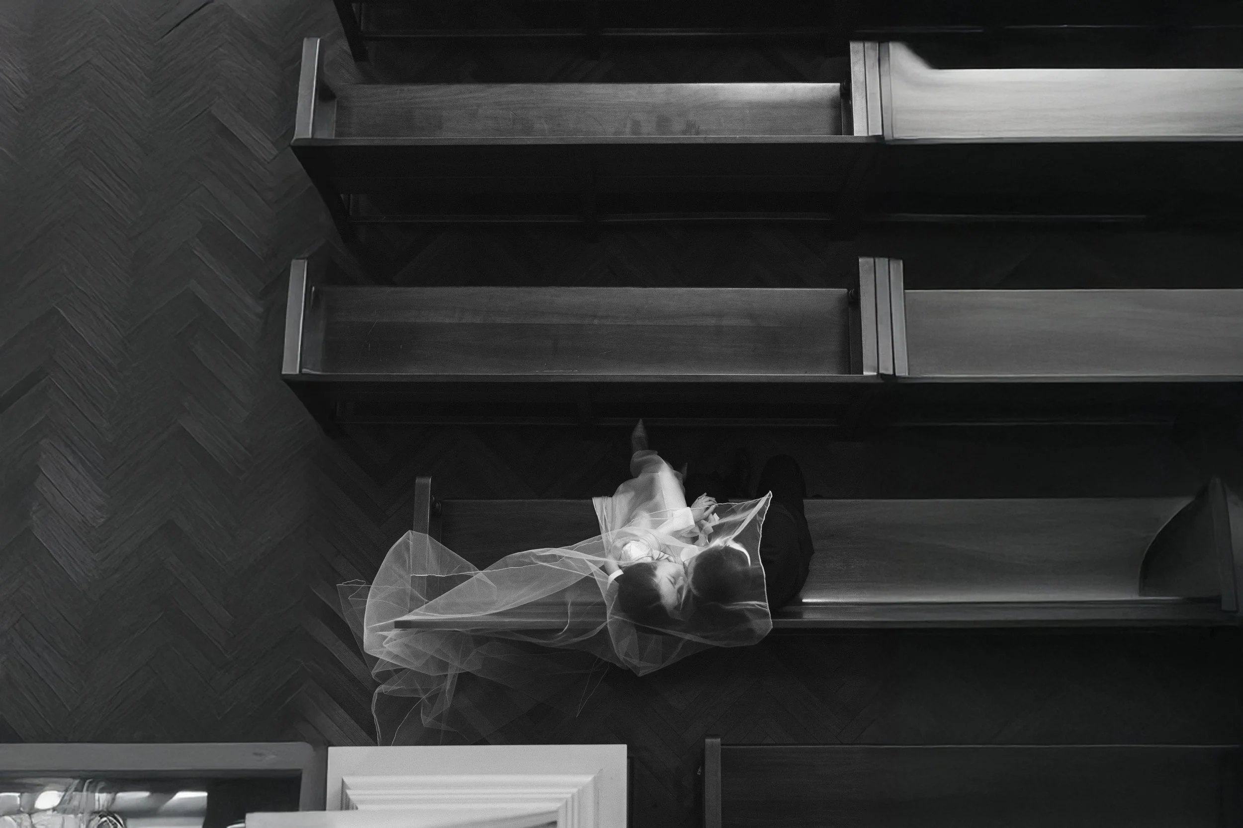 A girl wearing a veil is sitting alone on a wooden church bench, looking up, with an overhead view in black and white.