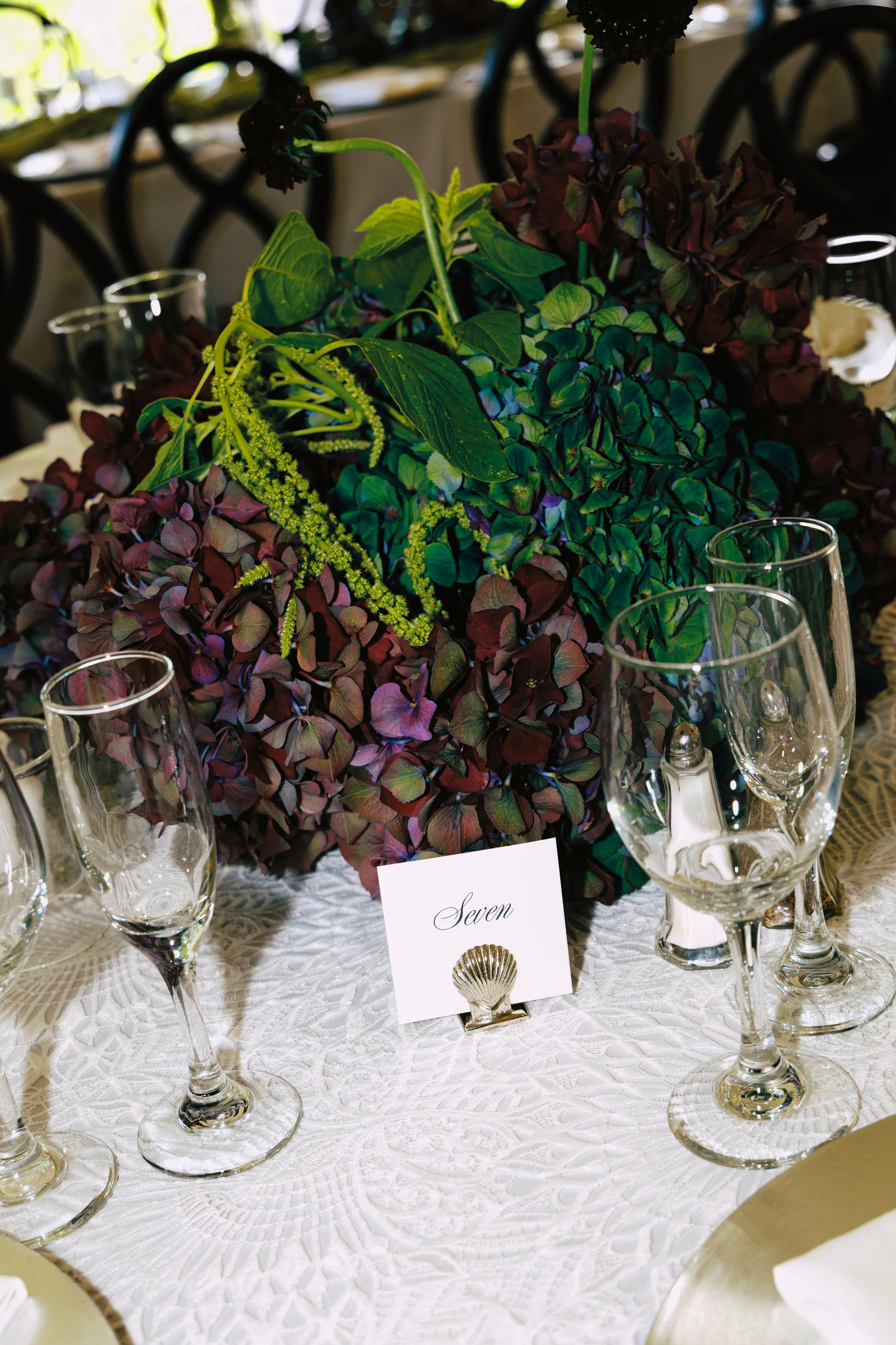 A table setting with a floral centerpiece of purple and green hydrangeas, a white tablecloth, and empty champagne glasses, with a table number card labeled 'Seven' in front of the arrangement.