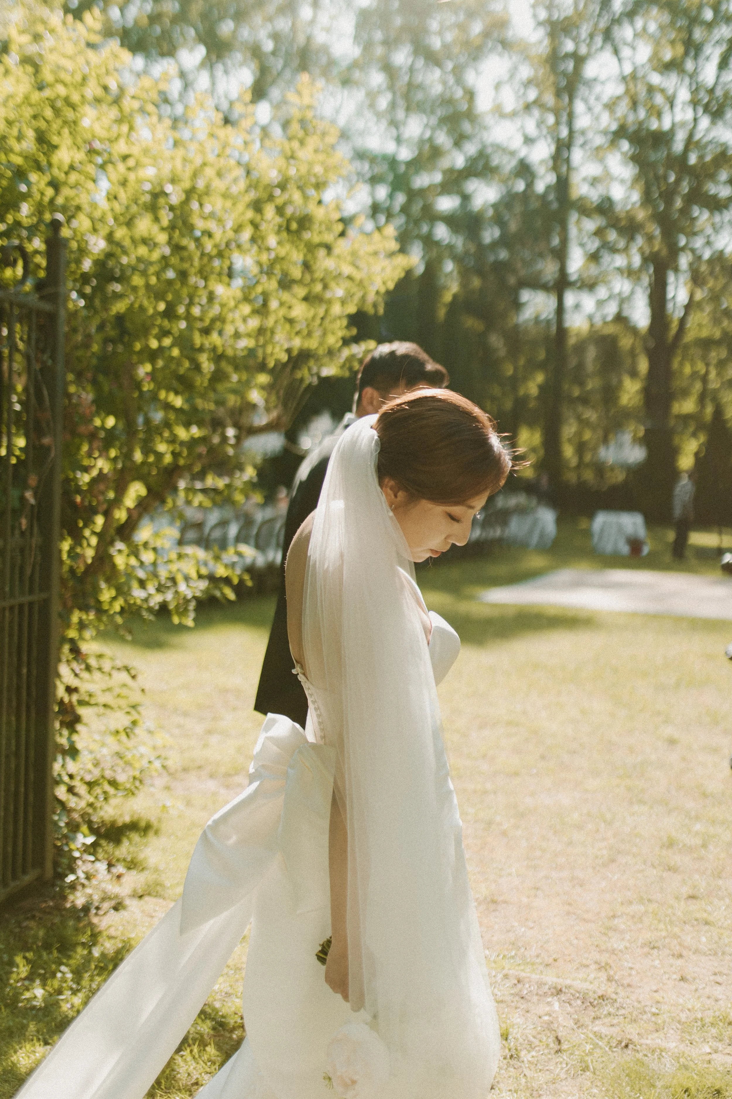 Bride in a white wedding dress and veil holding a bouquet, standing outdoors with a groom in a suit behind her, during daytime with sunlight filtering through trees.