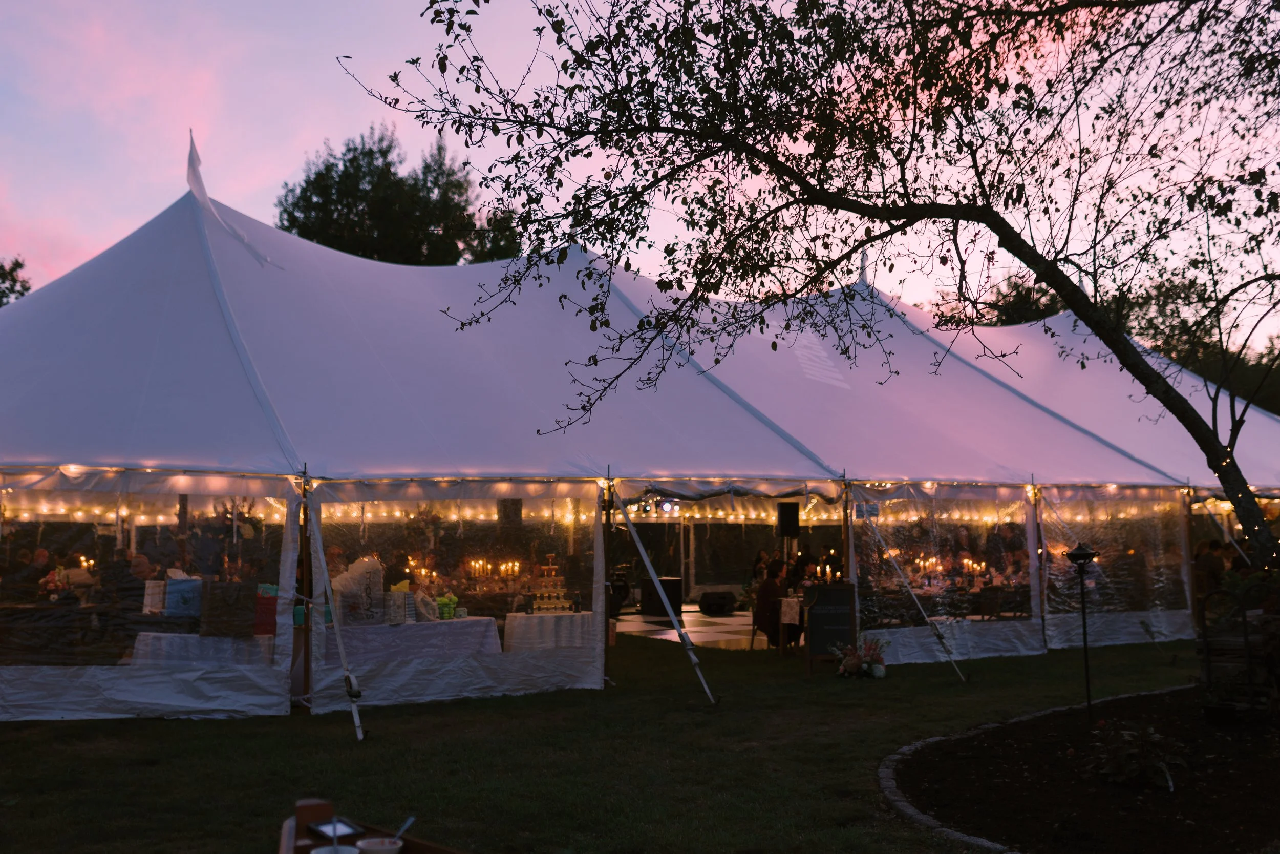 A large white event tent with string lights inside, set up outdoors on a grassy area during sunset, with trees and a pink sky in the background.
