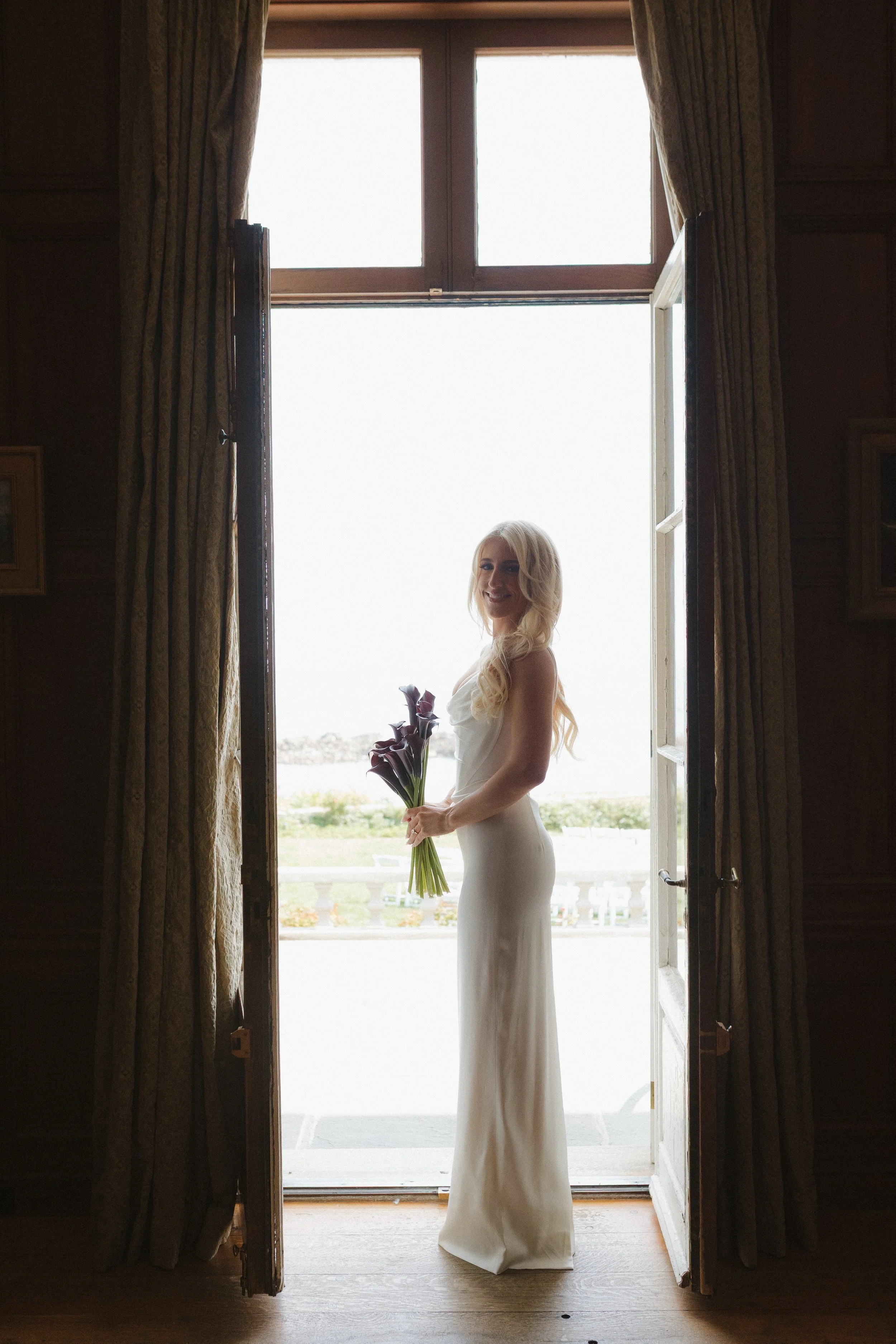 A woman in a white dress holding a bouquet of purple lilies standing in an open doorway, backlit by bright outdoor light, with wooden interior walls and curtains.