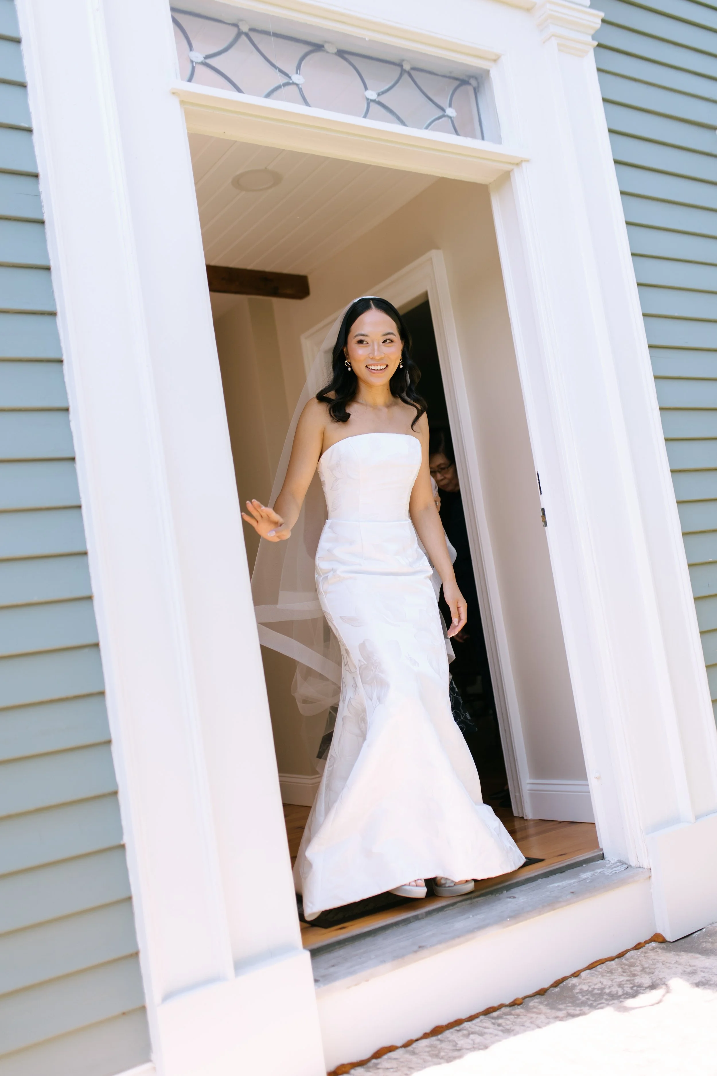 A woman in a strapless white wedding dress smiling as she greets someone at the doorway of a house.