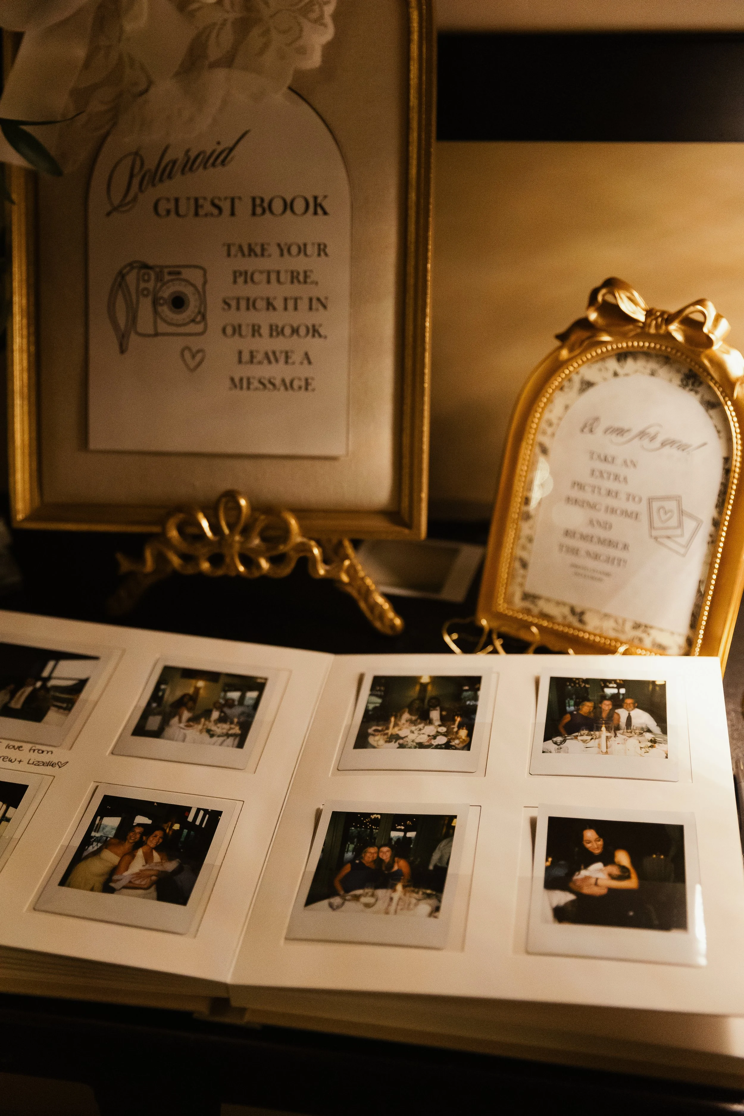 Polaroid guest book sign with instructions to take a picture and leave a message, and an accompanying smaller framed sign with similar instructions, alongside a photo album with several instant photos of people at a gathering placed on a table.