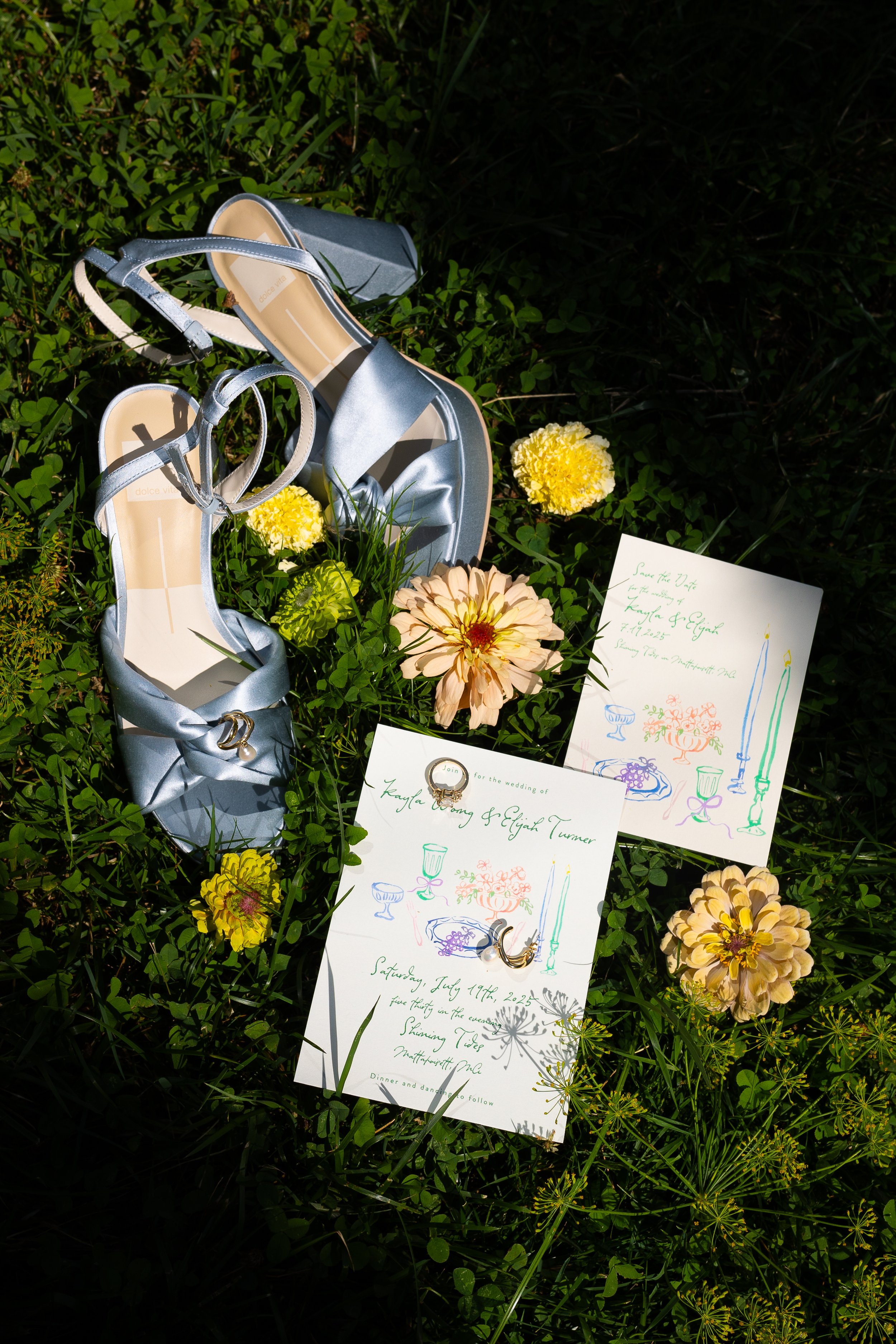 Silver high-heeled sandals, wedding invitation cards, flowers, and a ring placed on the grass.
