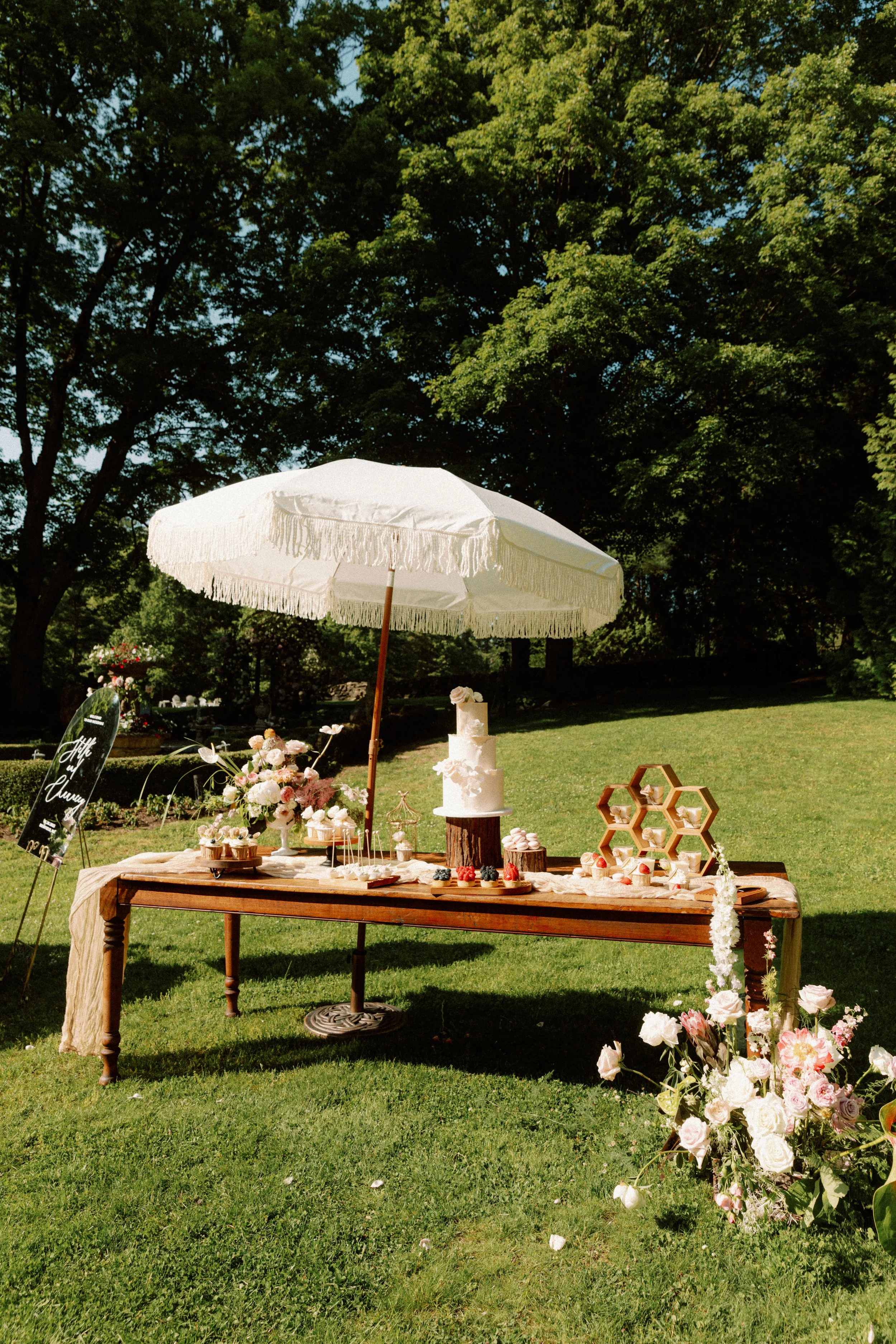 Outdoor dessert table with a white cake, cupcakes, and floral decorations under a large white umbrella on a grassy lawn surrounded by trees.