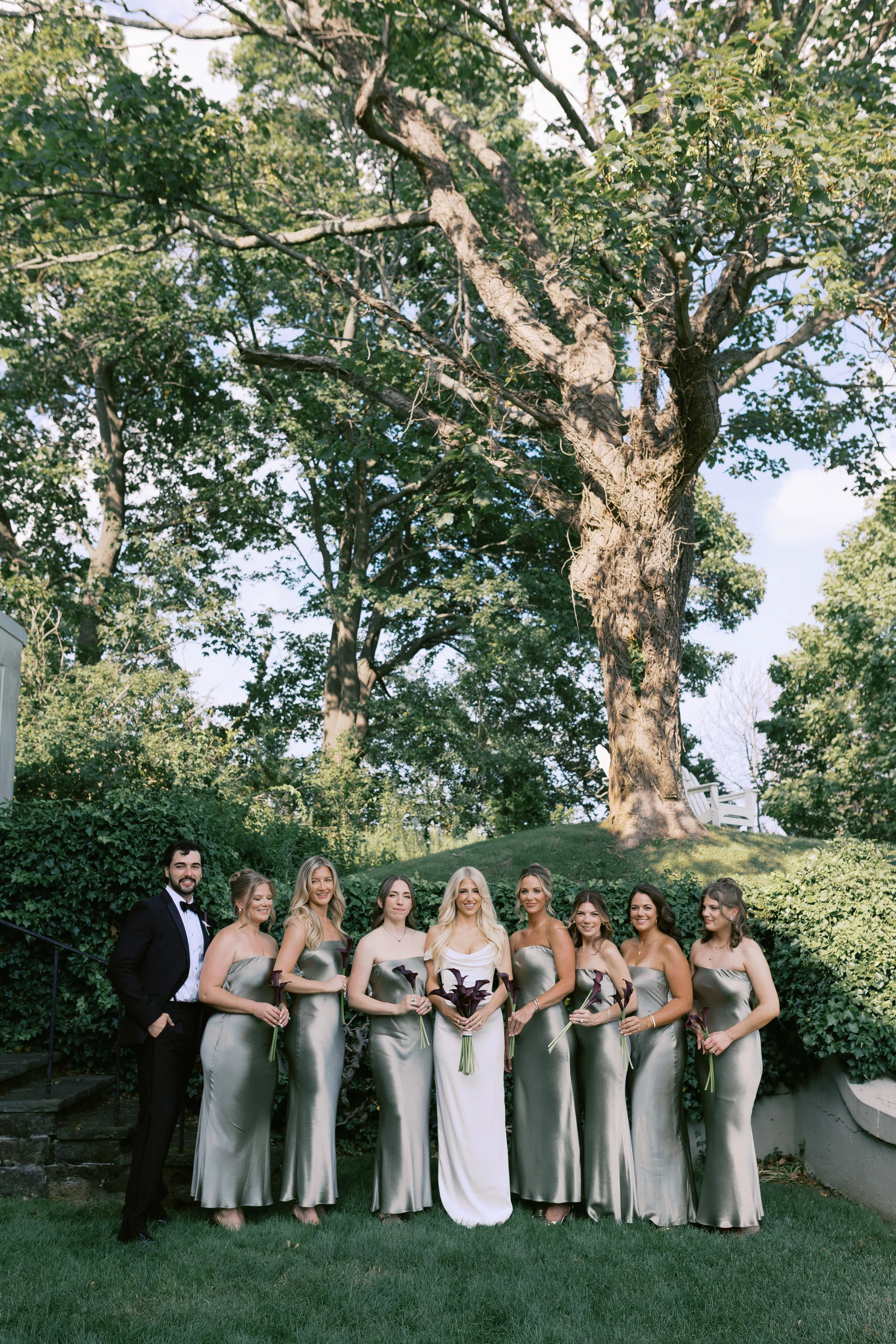 A bridal party standing outdoors on grass, with eight women in silver satin dresses and one man in a tuxedo, all holding flowers, under large trees.