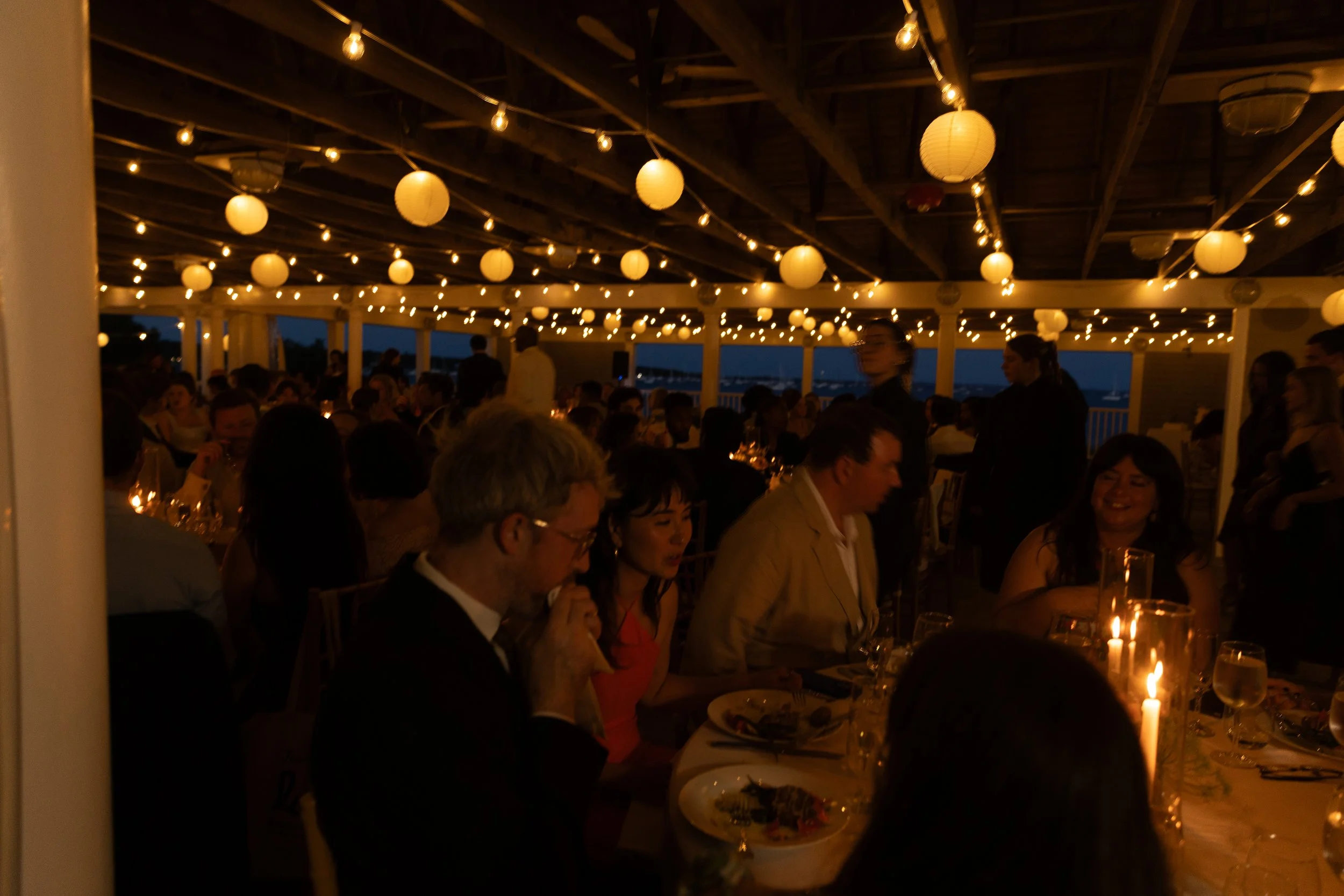 People enjoying dinner at a nighttime celebration with string lights and paper lanterns hanging from the ceiling of a decorated outdoor venue.