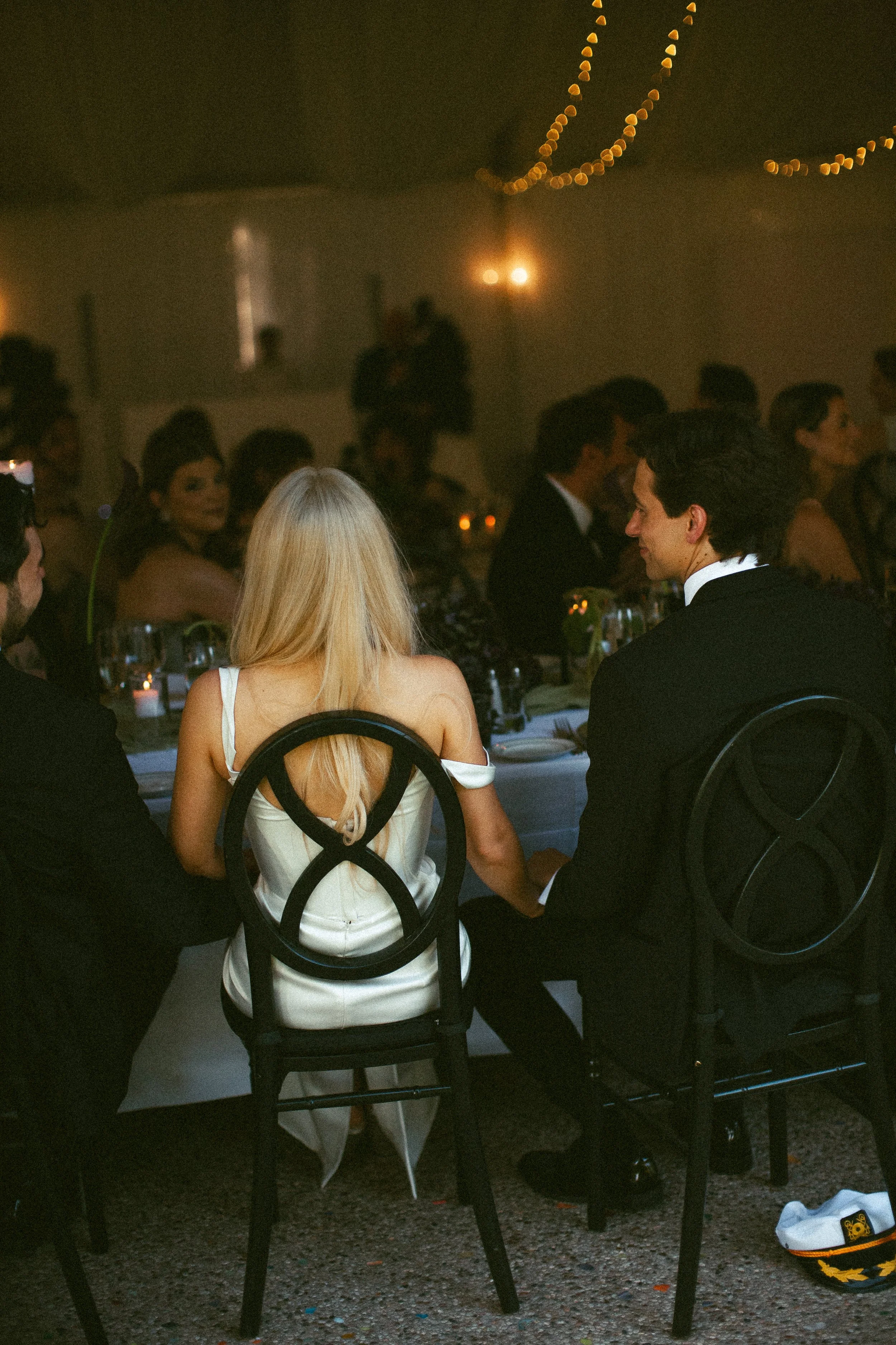 People seated at a formal dinner event; focus on a woman with long blonde hair in a white dress and a man in a tuxedo, sitting at a table with candles and tableware, under hanging string lights in a dimly lit room.