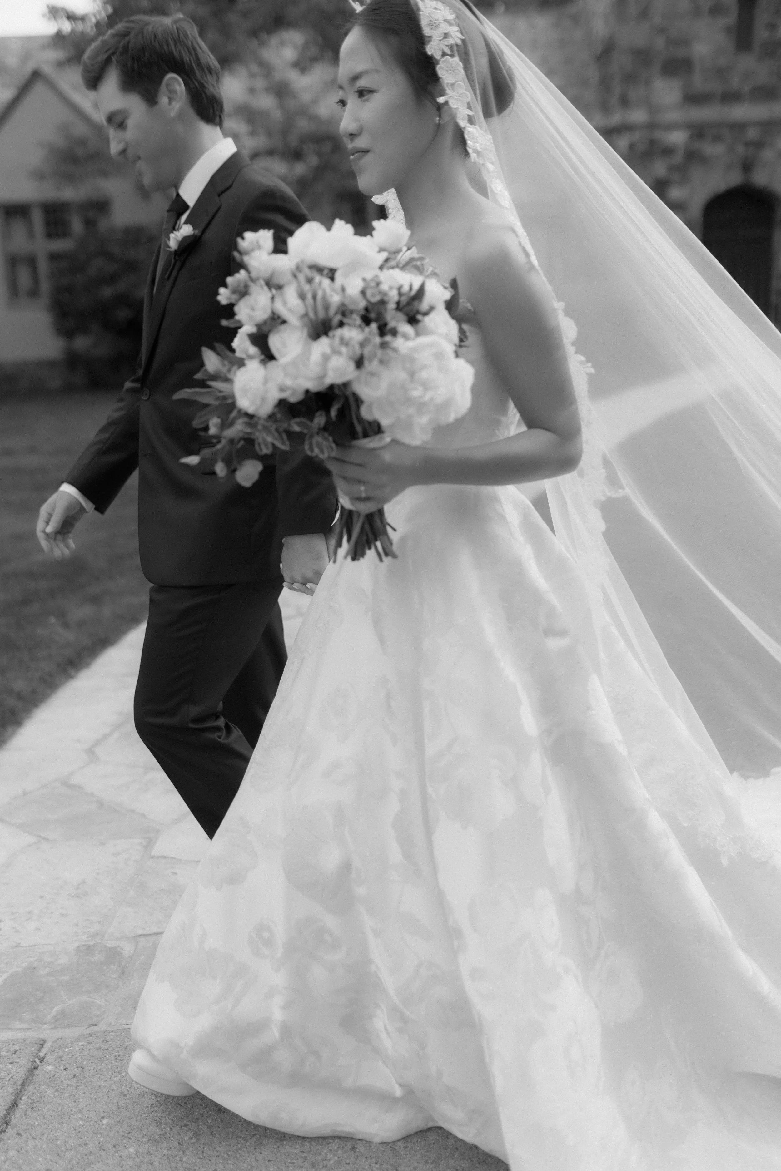 A bride and groom walking hand in hand outdoors, the bride holding a bouquet of flowers. The bride is wearing a wedding dress with a long veil, and the groom is dressed in a suit with a boutonniere. The photo is in black and white.