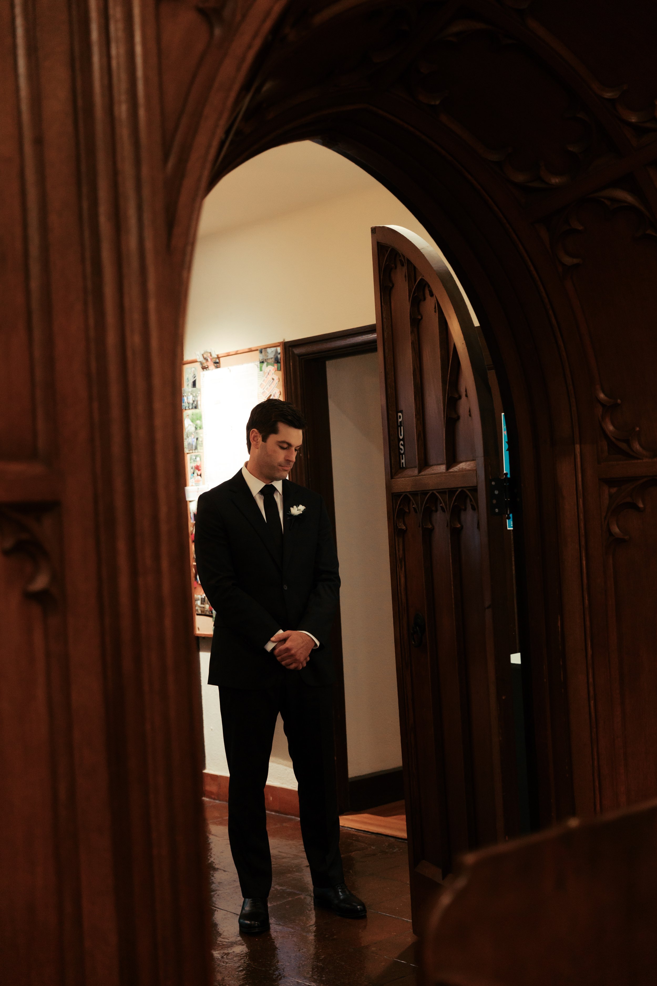 A man dressed in a black suit with a white shirt and tie, standing with hands clasped, inside a room with dark wooden furniture, possibly preparing for a formal event or ceremony.