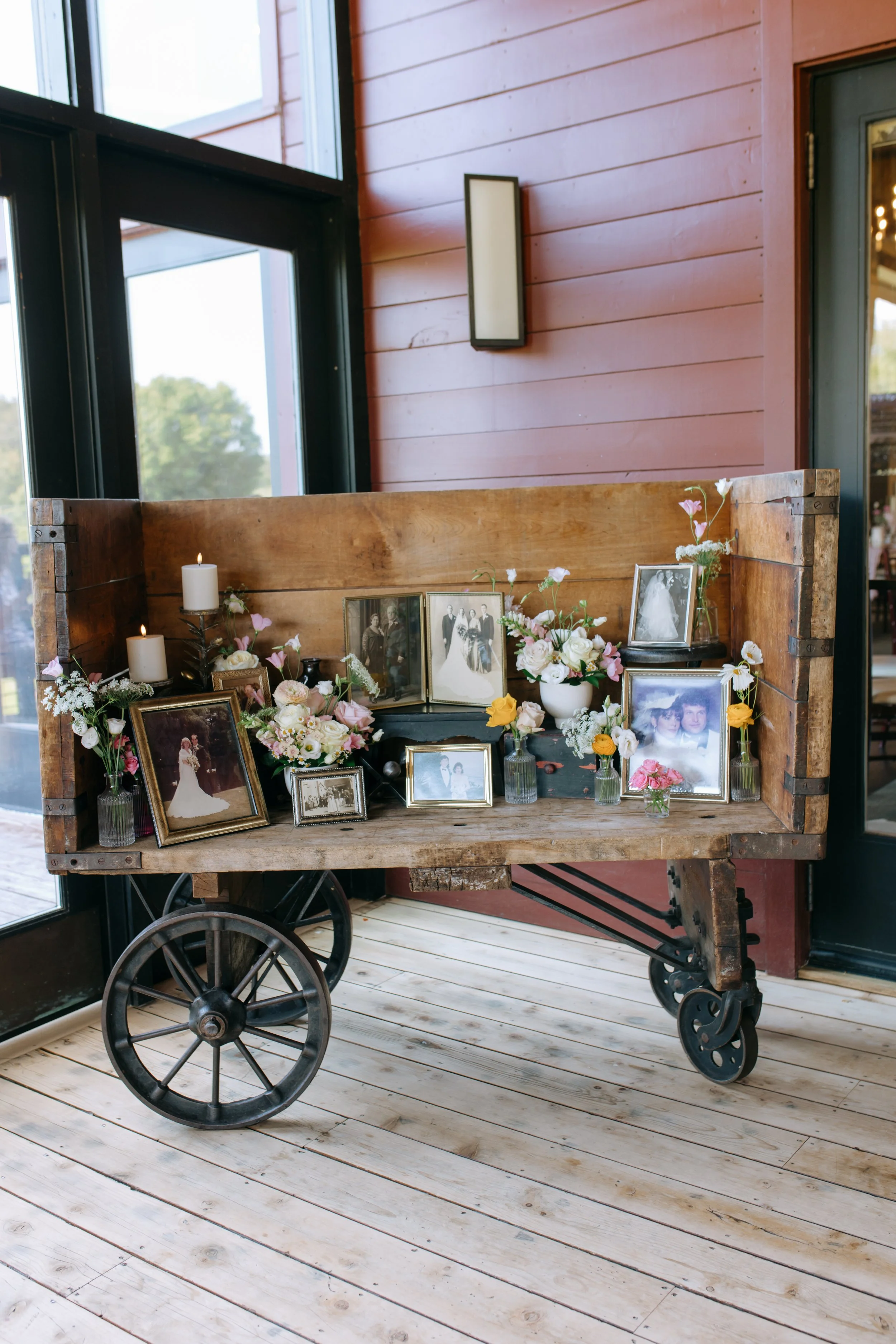 A vintage wooden cart with black metal wheels and a wooden back panel, decorated with framed photographs and flower arrangements at a corner of a room with large windows and wood-paneled walls.