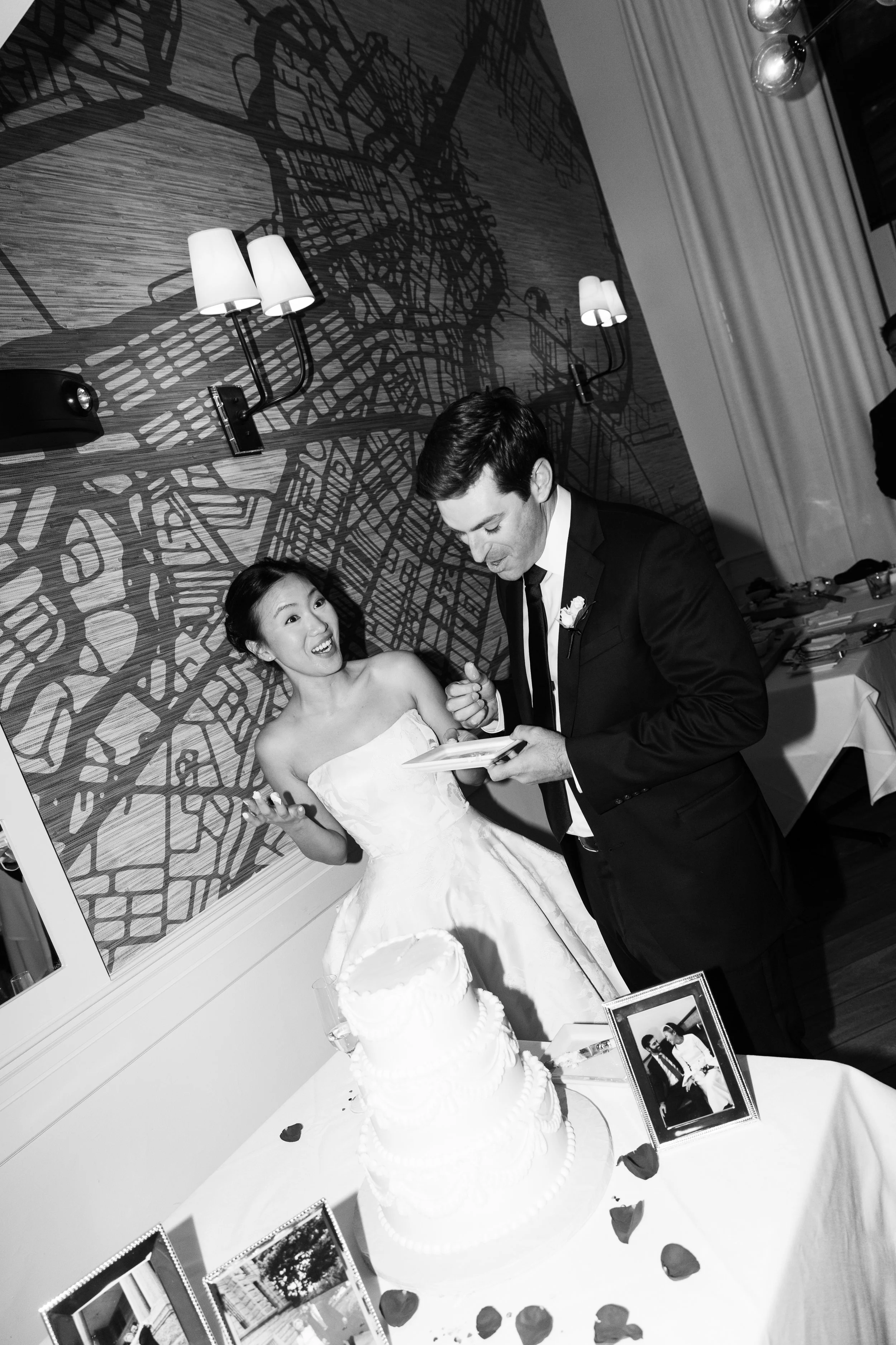 Black and white photo of a newlywed couple at their wedding reception, exchanging a moment of joy near a wedding cake with framed photos on the table, with decorative wall art in the background.