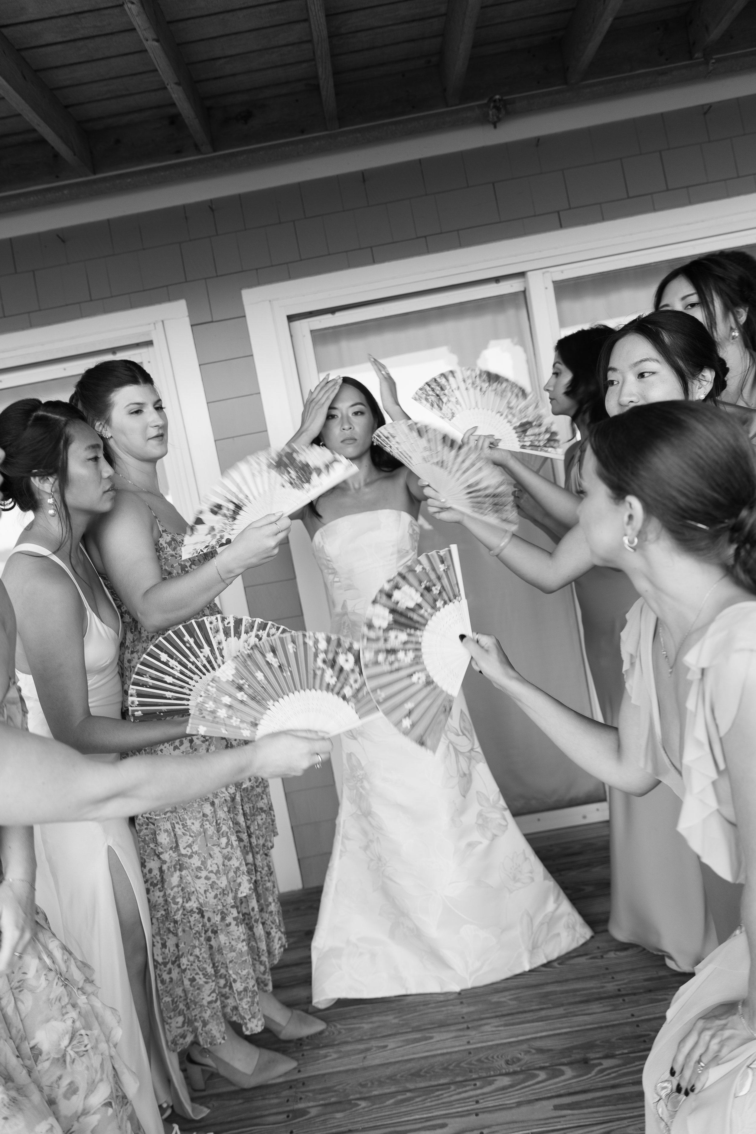 A bride surrounded by her bridesmaids holding decorative fans, in a joyful moment during a wedding preparation.