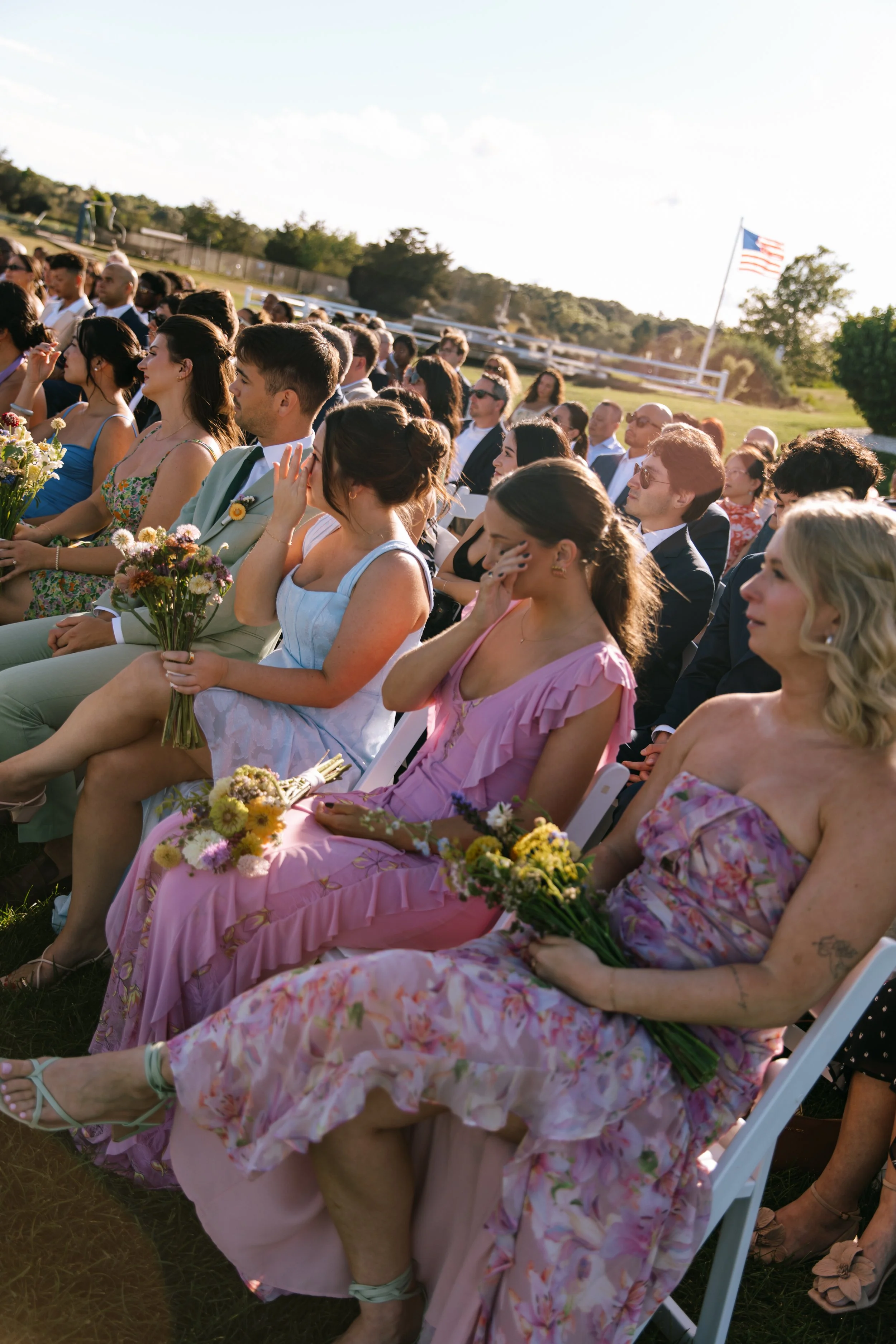 People attending an outdoor wedding ceremony, seated on white chairs, holding bouquets, with a flagpole and American flag in the background.
