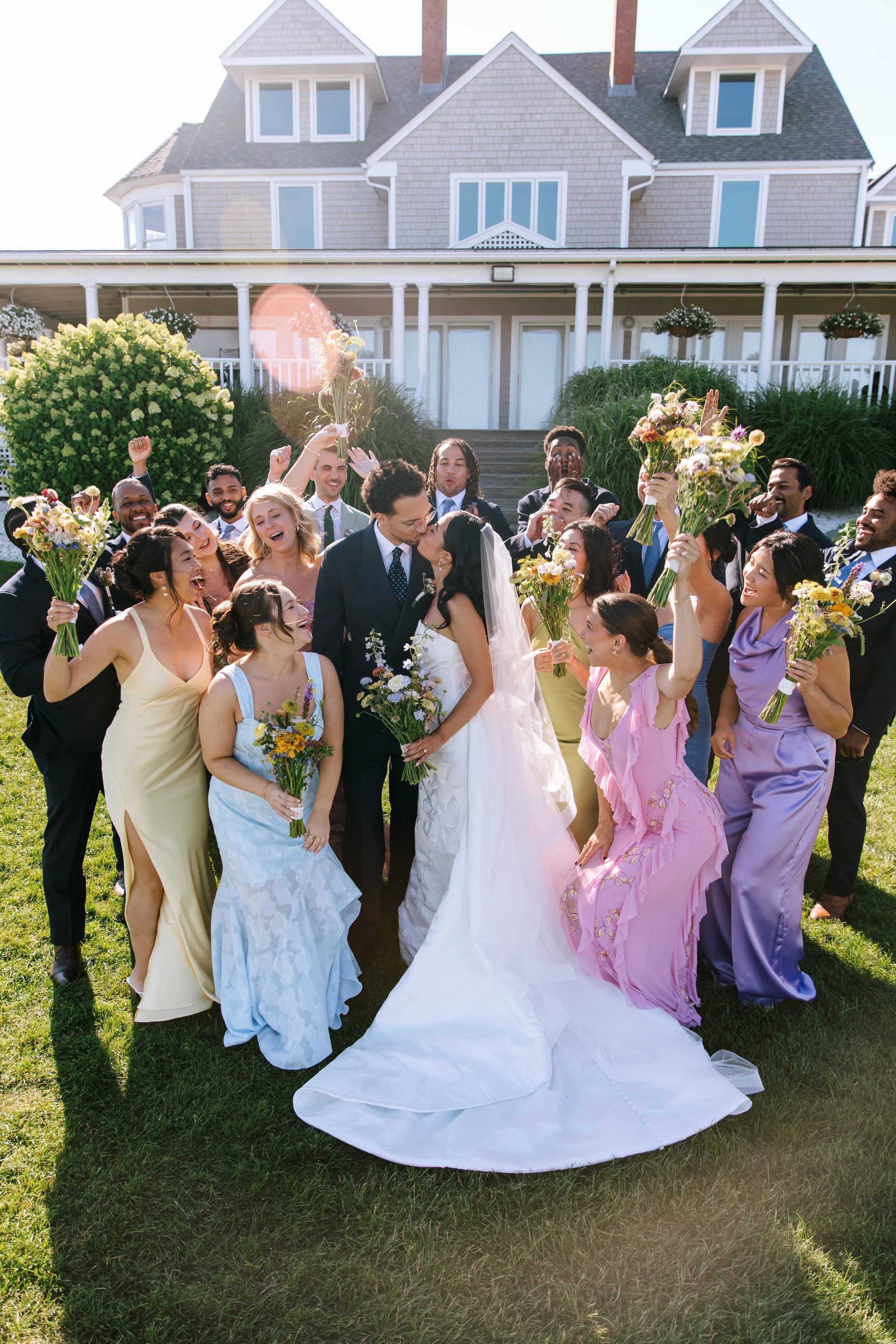 A diverse group of people celebrating at a wedding outdoors, with two people in the center kissing, surrounded by friends holding bouquets and cheering.