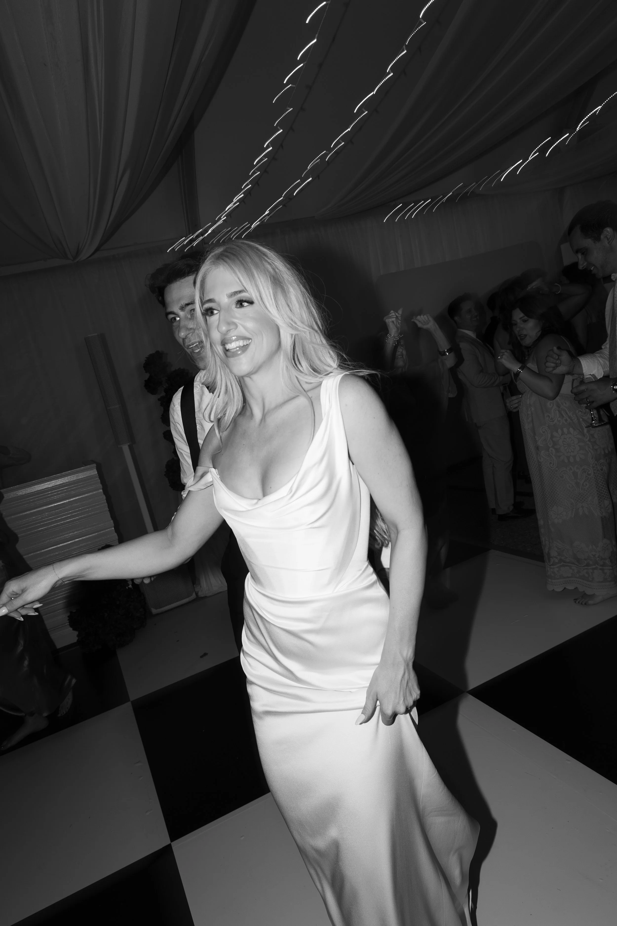 A woman in a white satin gown dancing at a celebration or wedding reception, smiling and enjoying the moment, with other guests dancing in the background.