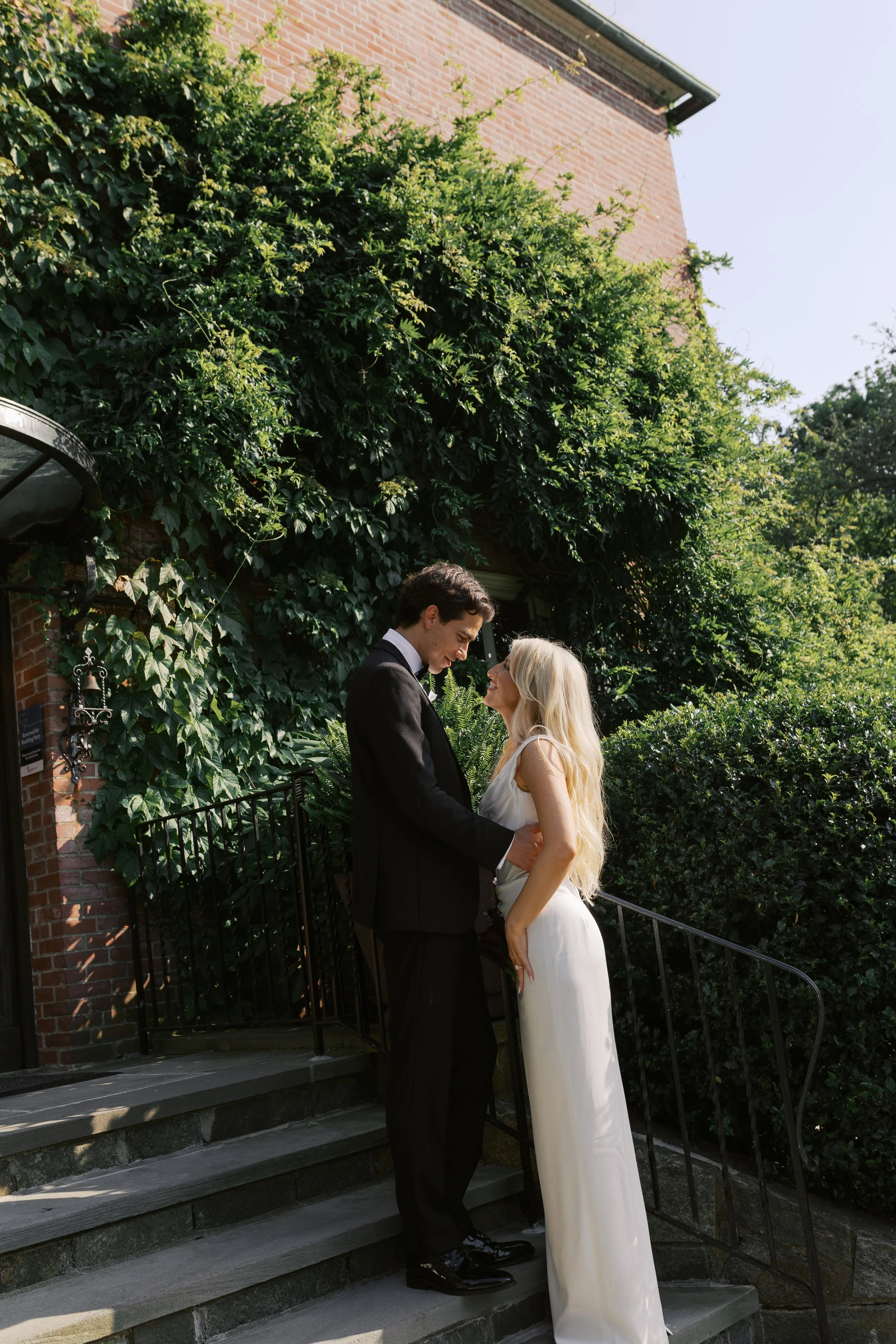 A couple in wedding attire standing on outdoor steps, smiling at each other, with lush green foliage and a brick building in the background.