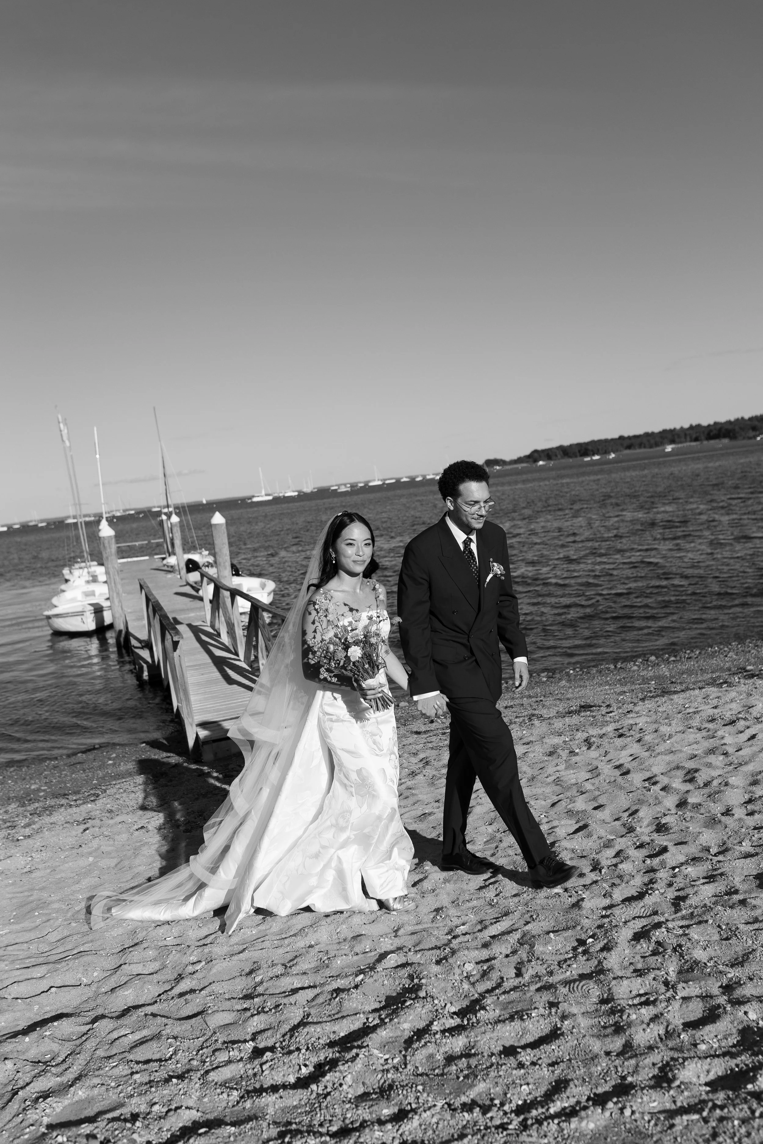 A newlywed couple in wedding attire walking along a sandy beach near a water dock with boats in the background, captured in black and white.
