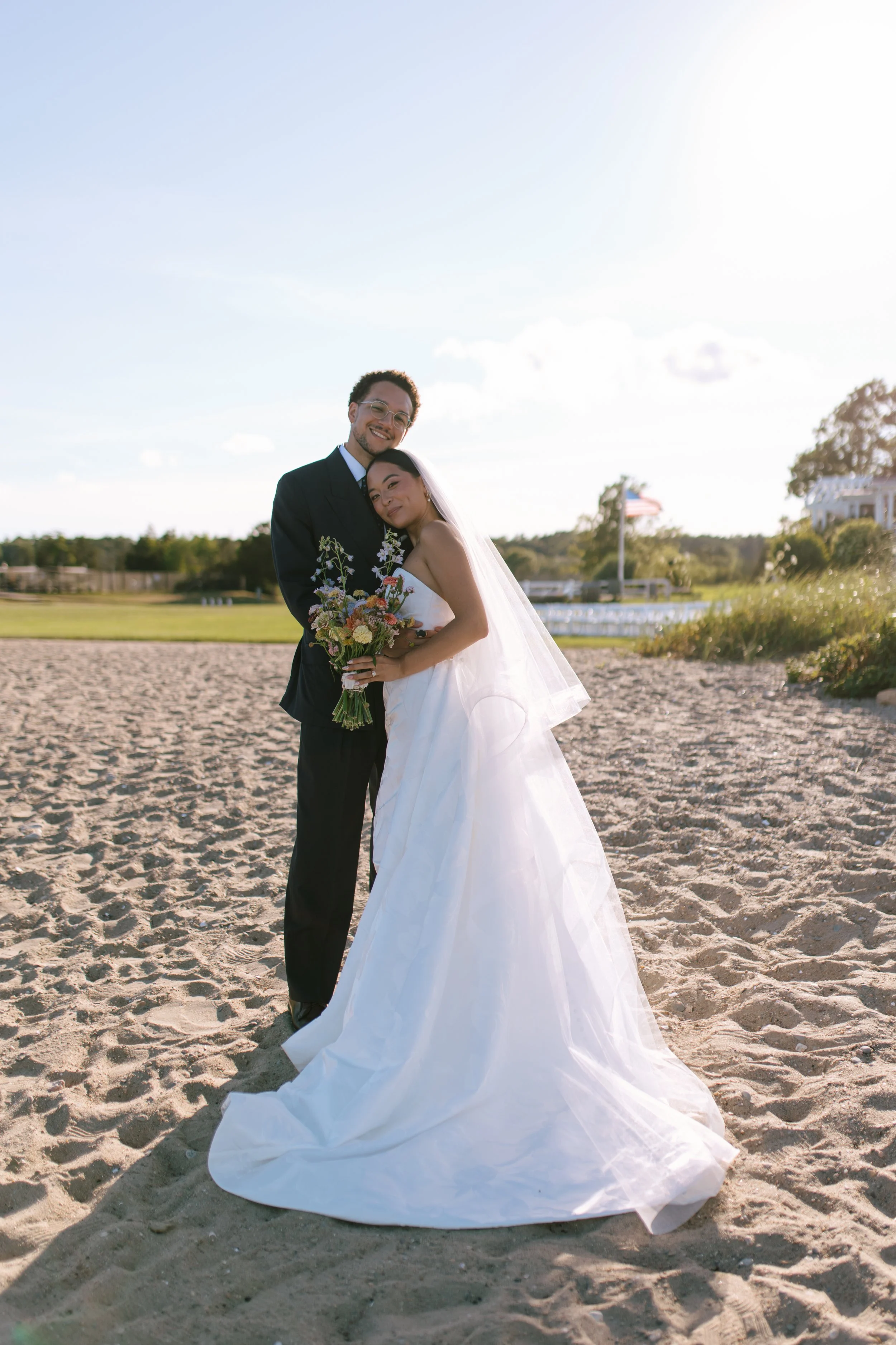 A newlywed couple embracing on a sandy beach, with the bride holding a bouquet of flowers and both smiling, under a bright sky during sunset or late afternoon.