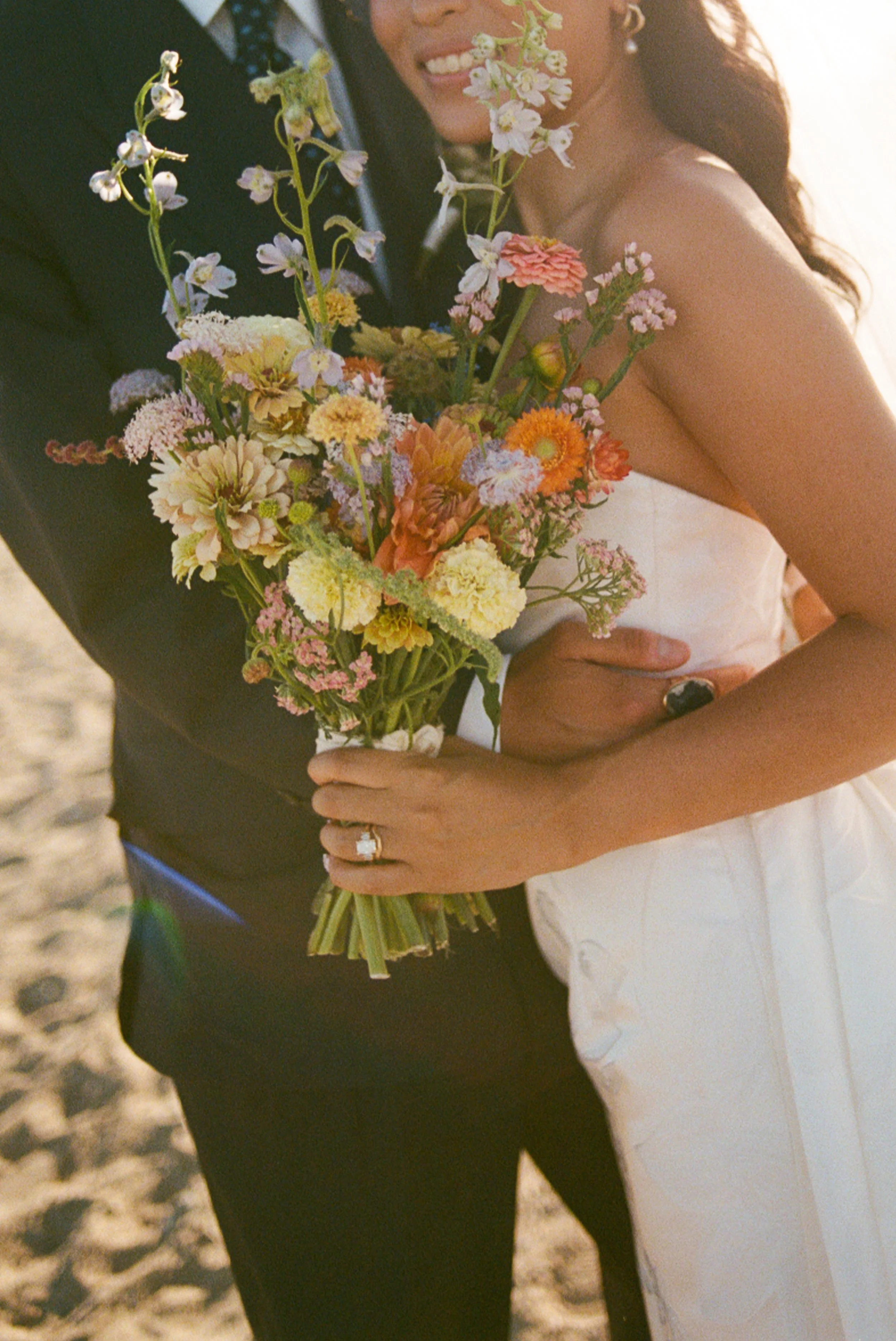 A couple dressed in wedding attire embracing outdoors, with the woman holding a bouquet of colorful flowers and smiling.