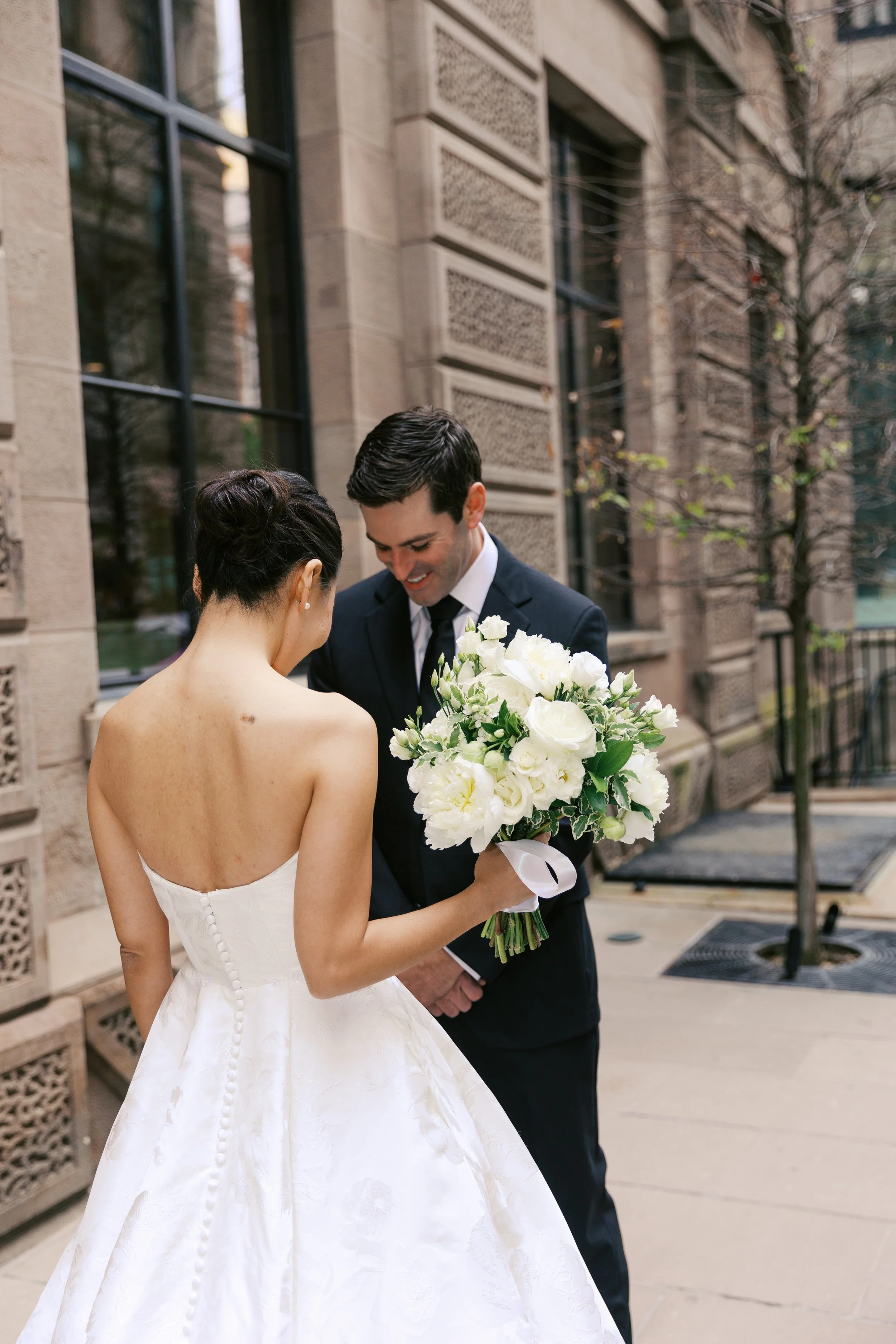 A bride in a white wedding dress holding a bouquet of white flowers, smiling at a groom in a dark suit outside a brick building.