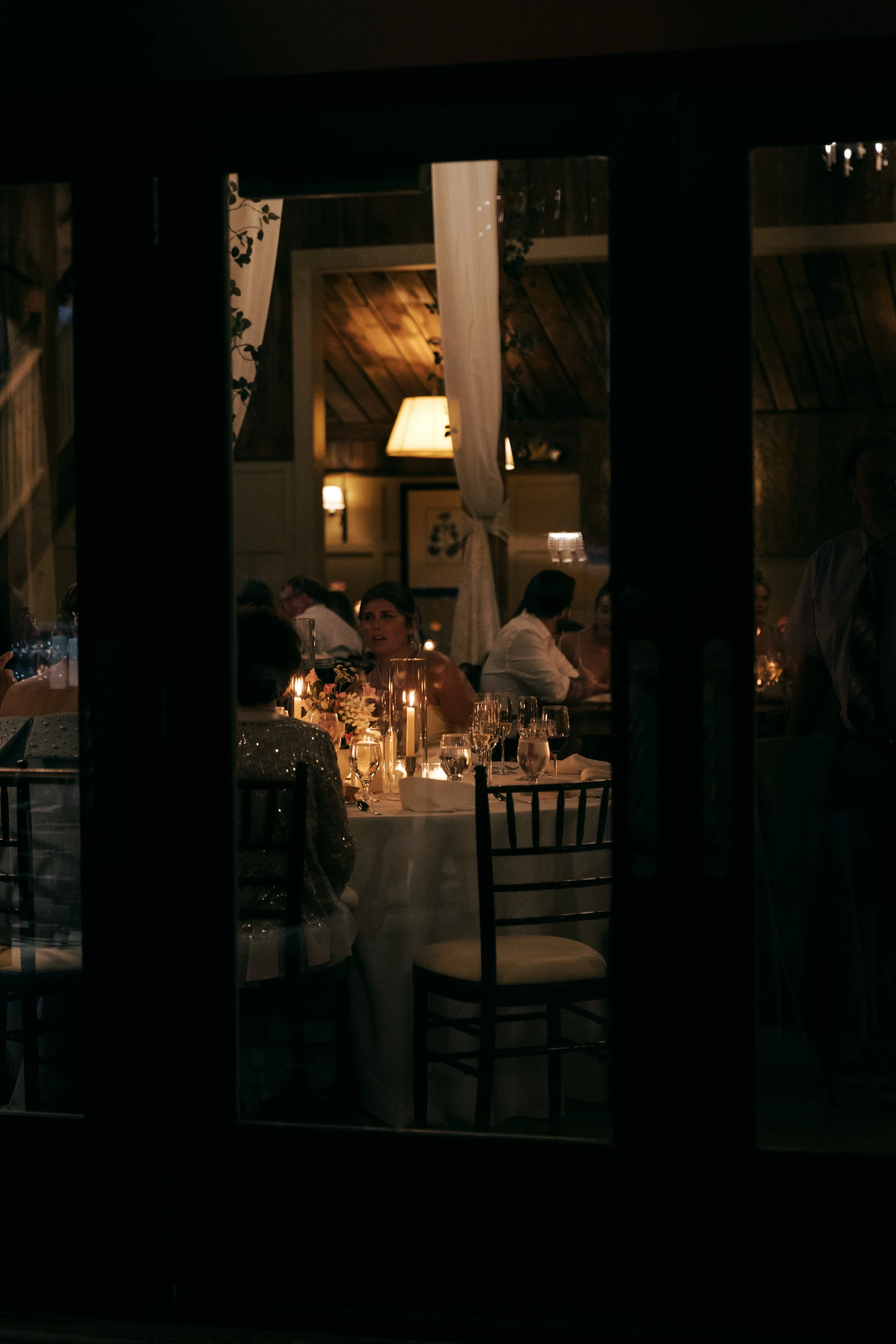 People dining at a restaurant table with candles, viewed through a glass window or door at night.
