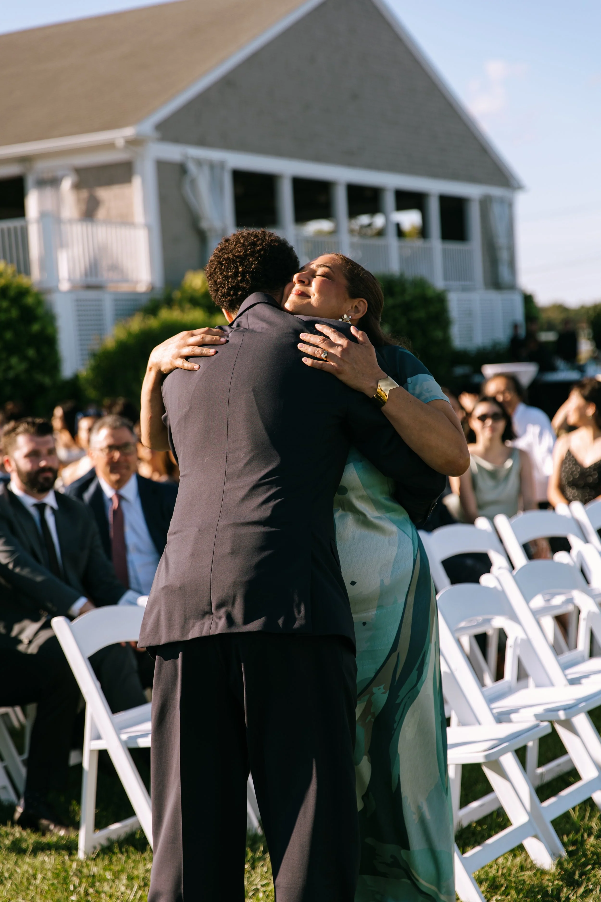 A woman embraces a man at an outdoor wedding ceremony with guests seated in white chairs behind them.