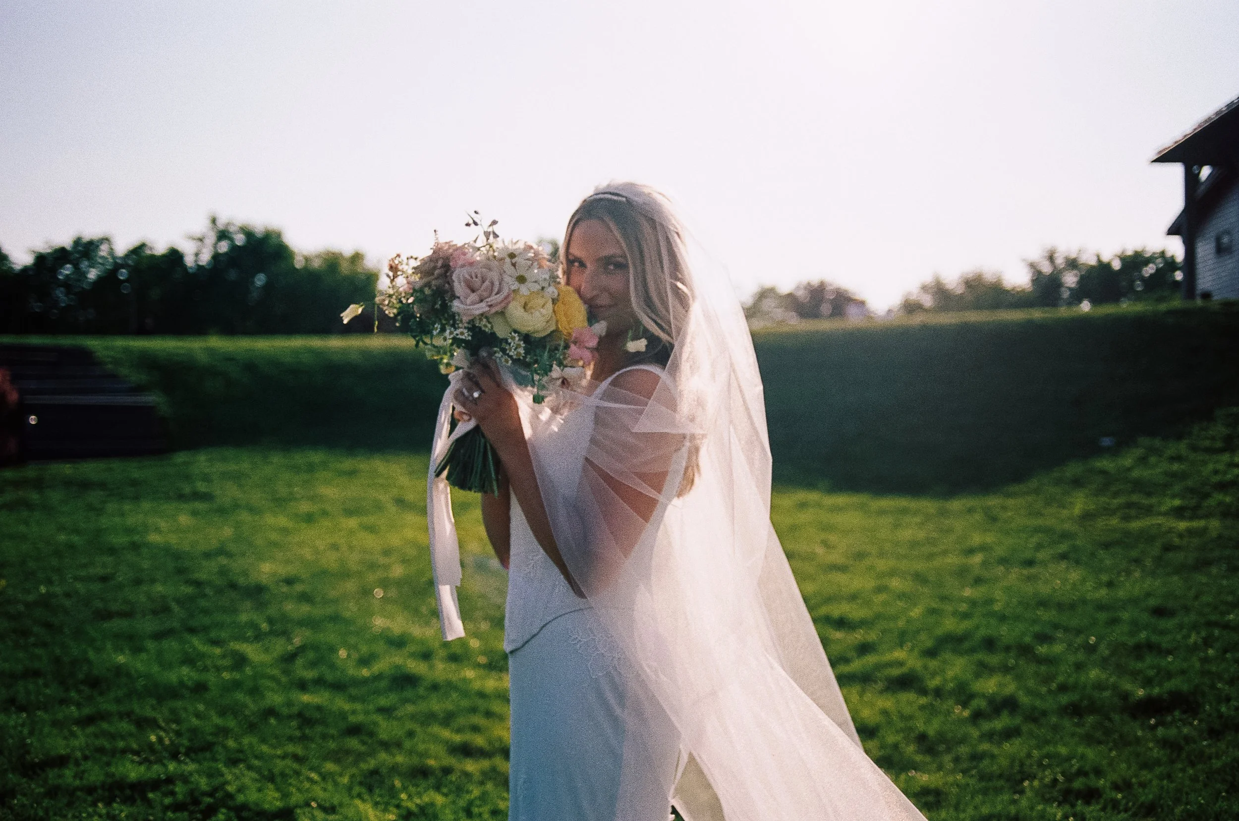 A bride in a white dress and veil holding a bouquet of flowers outdoors during sunset, with a grassy field and house in the background.