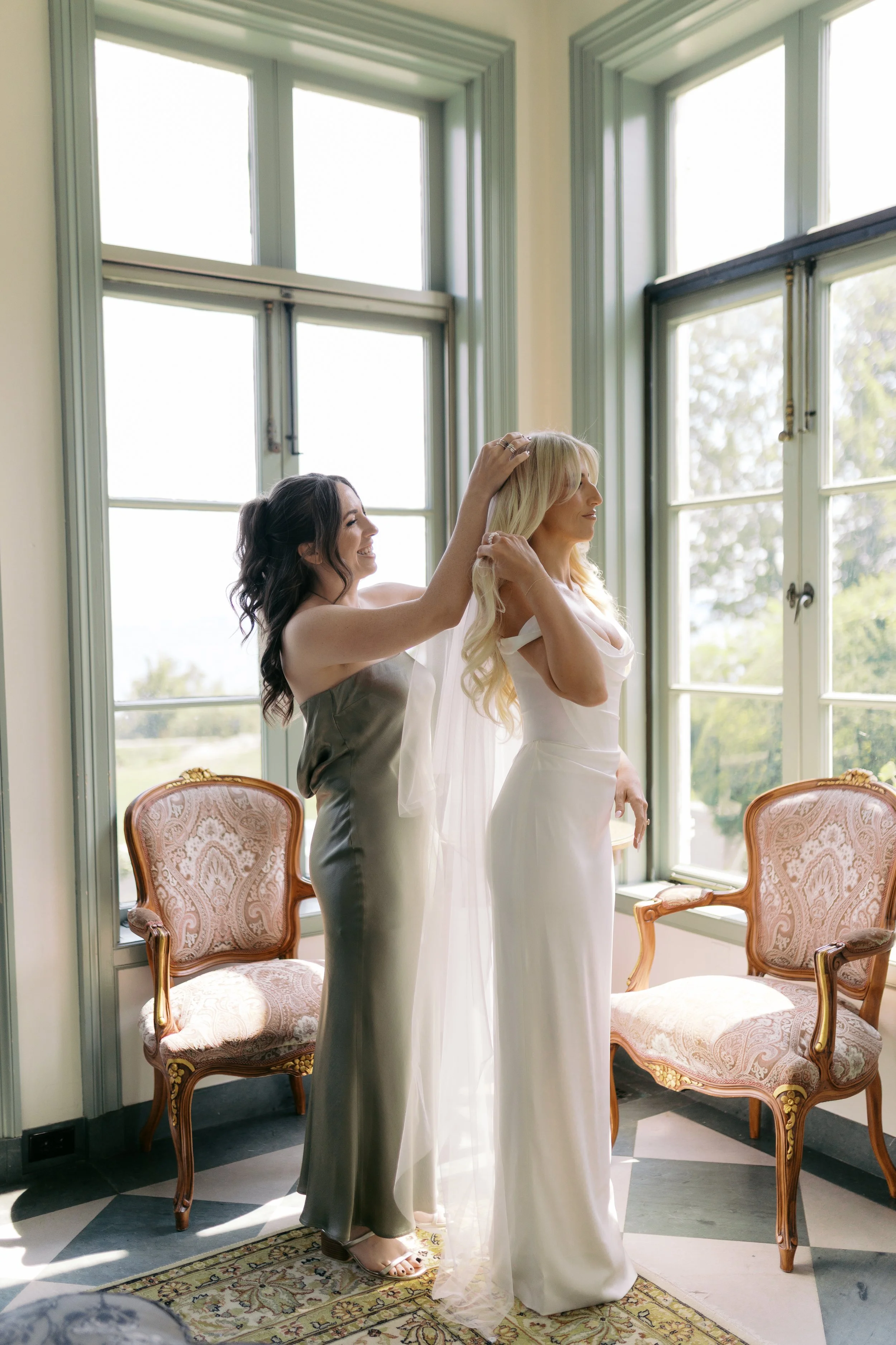 A bride in a white gown is having her hair adjusted by a woman in a satin dress, in a room with large windows and vintage chairs.