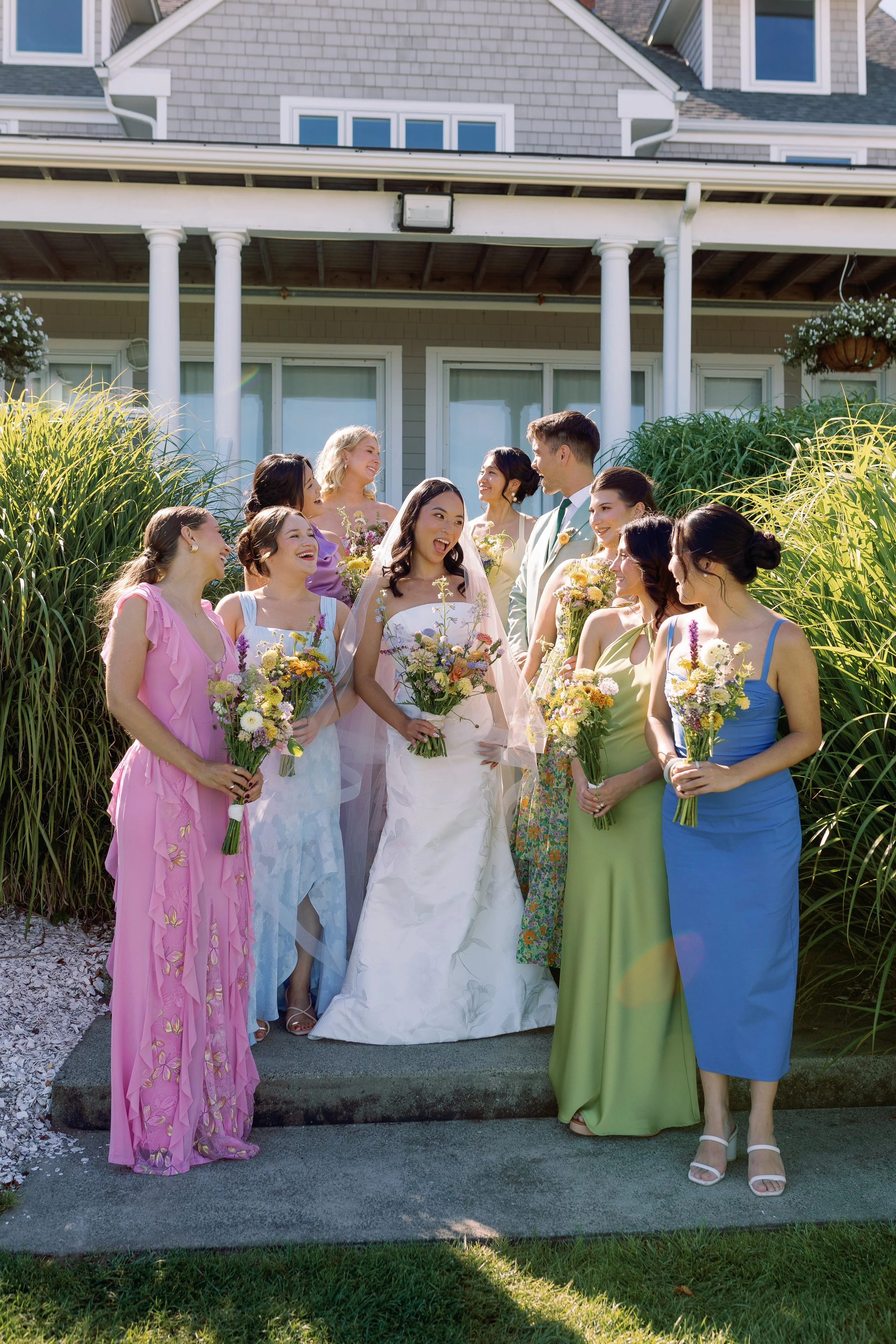 A wedding party with the bride in a white dress and bridesmaids in colorful dresses, standing outdoors in front of a house, all holding bouquets of flowers and enjoying a joyful moment.
