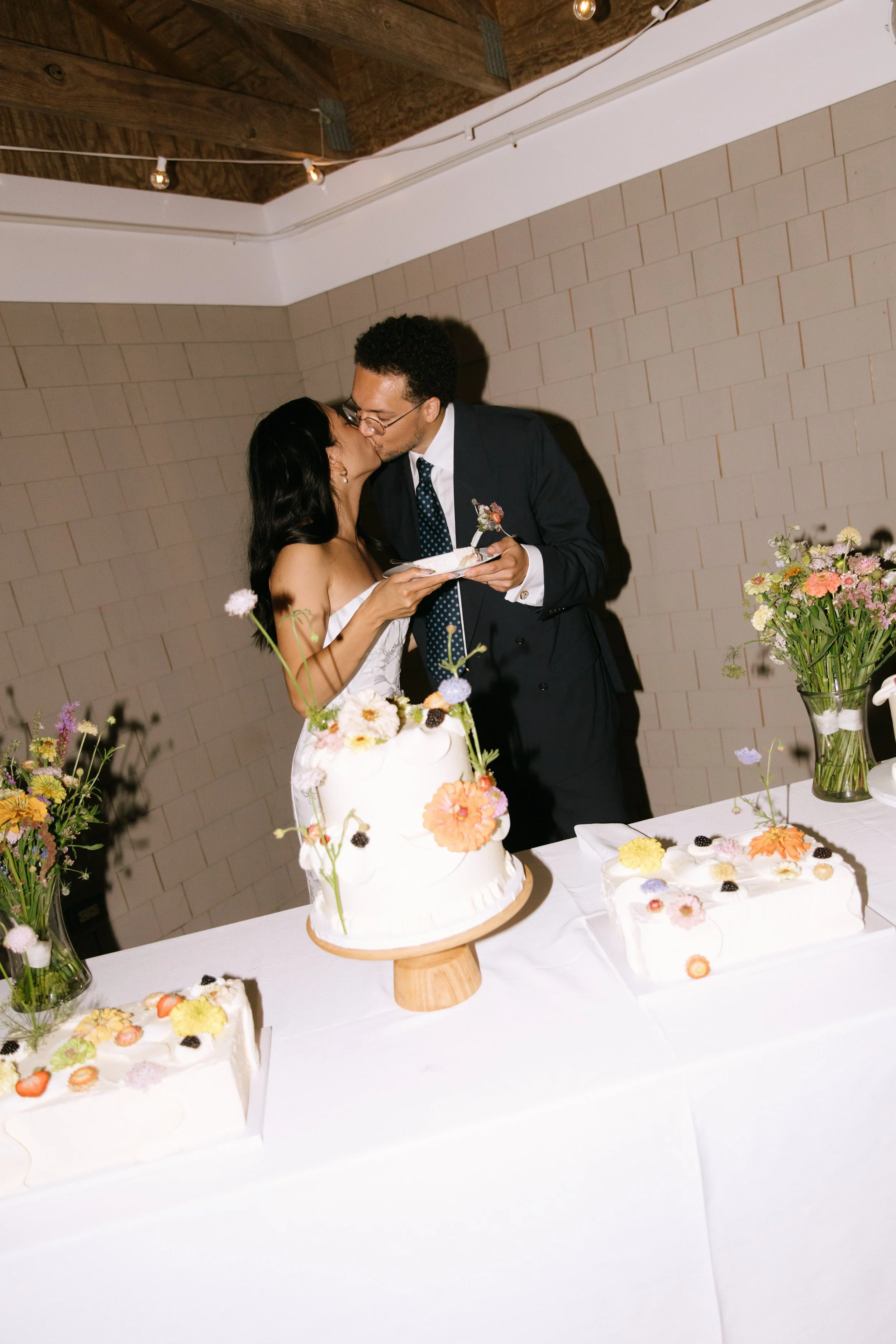 A newly married couple sharing a kiss during their wedding reception, with a wedding cake decorated with flowers on a table in front of them.