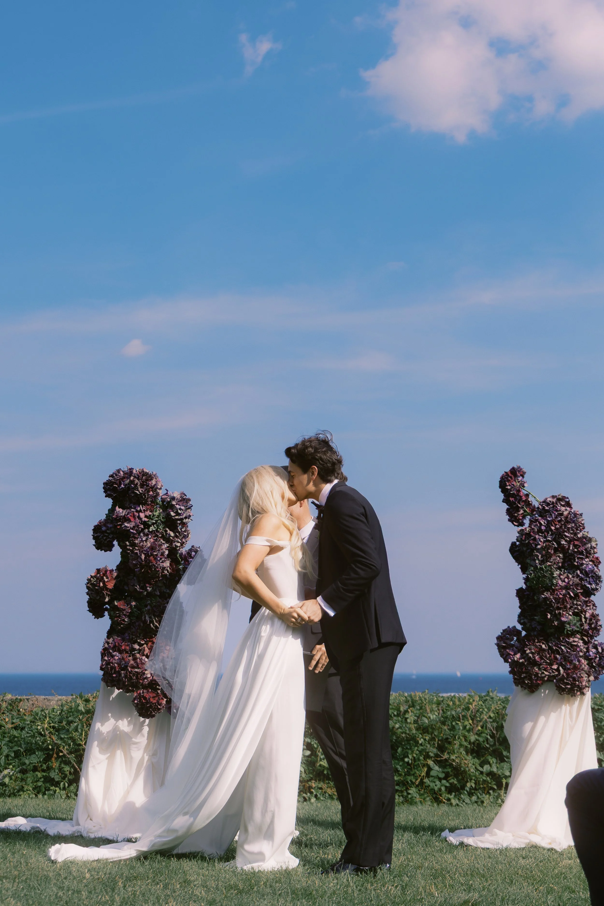 A bride and groom kissing at their outdoor wedding ceremony with floral arrangements and a blue sky background.