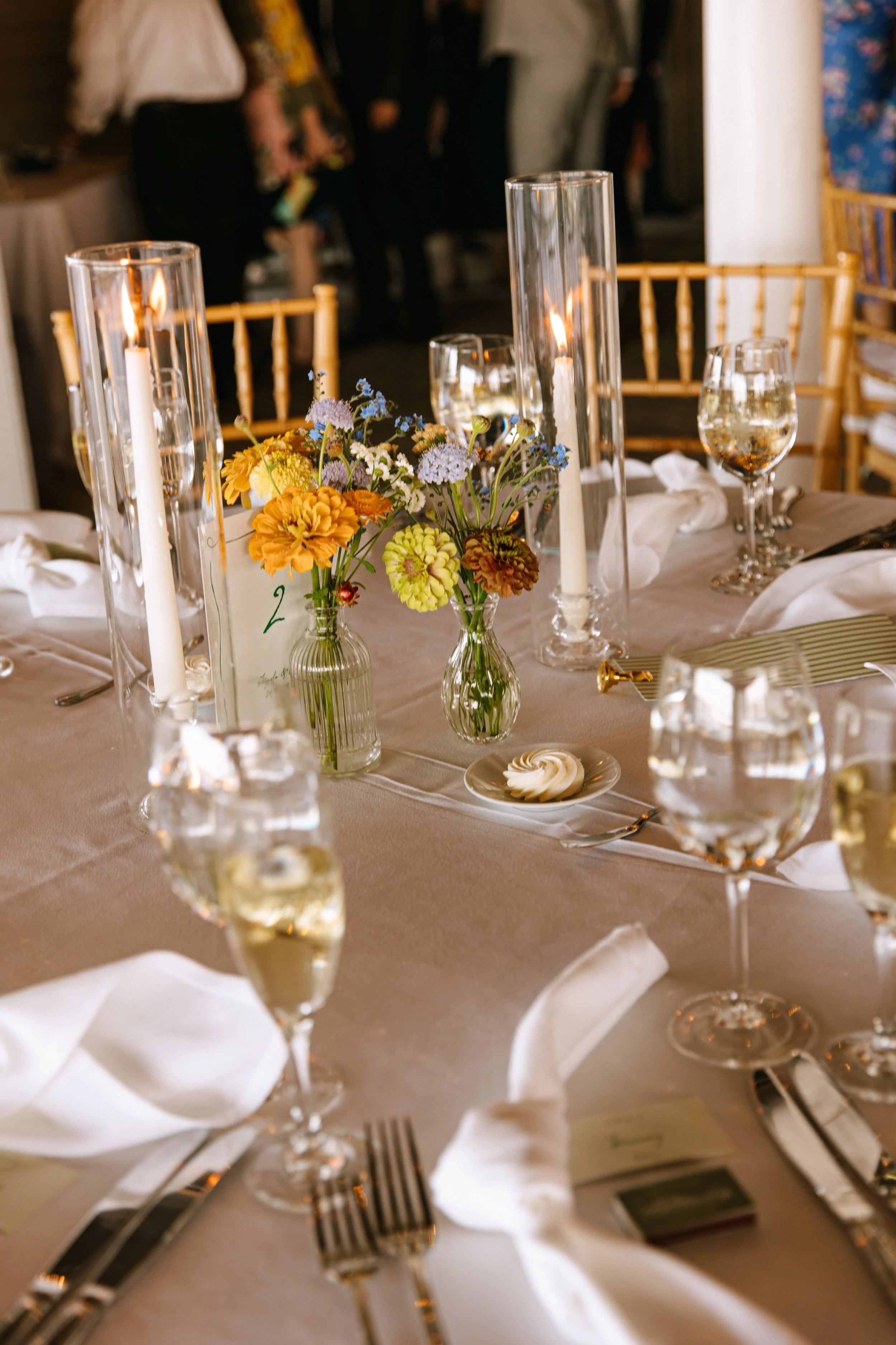 Elegant dining table with floral centerpiece, lit candles in tall glass holders, and place settings including glasses of wine or water, silverware, and white napkins, at a formal event or wedding reception.