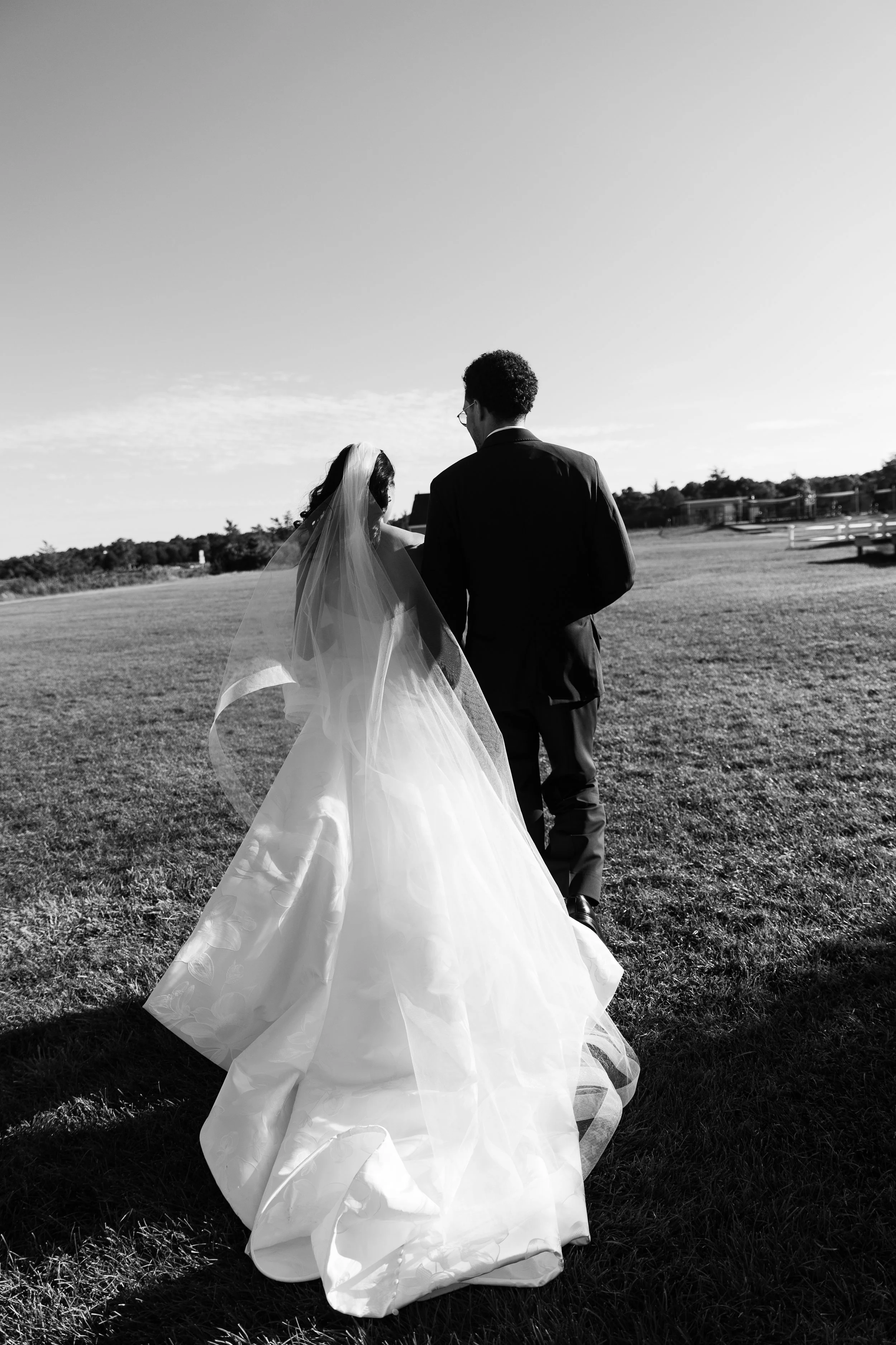 A black and white photo of a bride and groom walking together on a grassy field during sunset, with a clear sky and trees in the background.