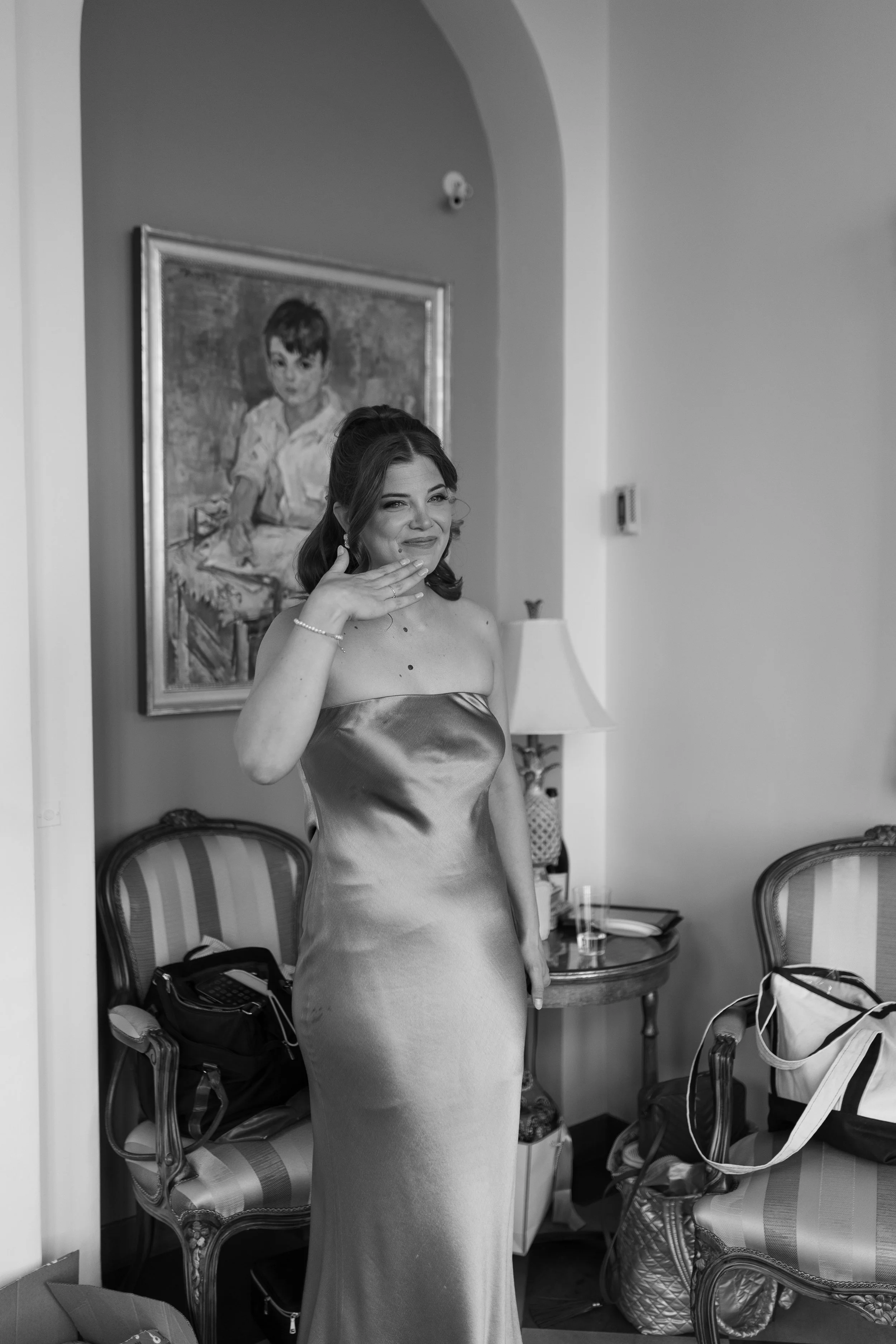 A woman in a strapless satin gown standing in a room, smiling and touching her face, with a portrait of a young boy on the wall behind her.
