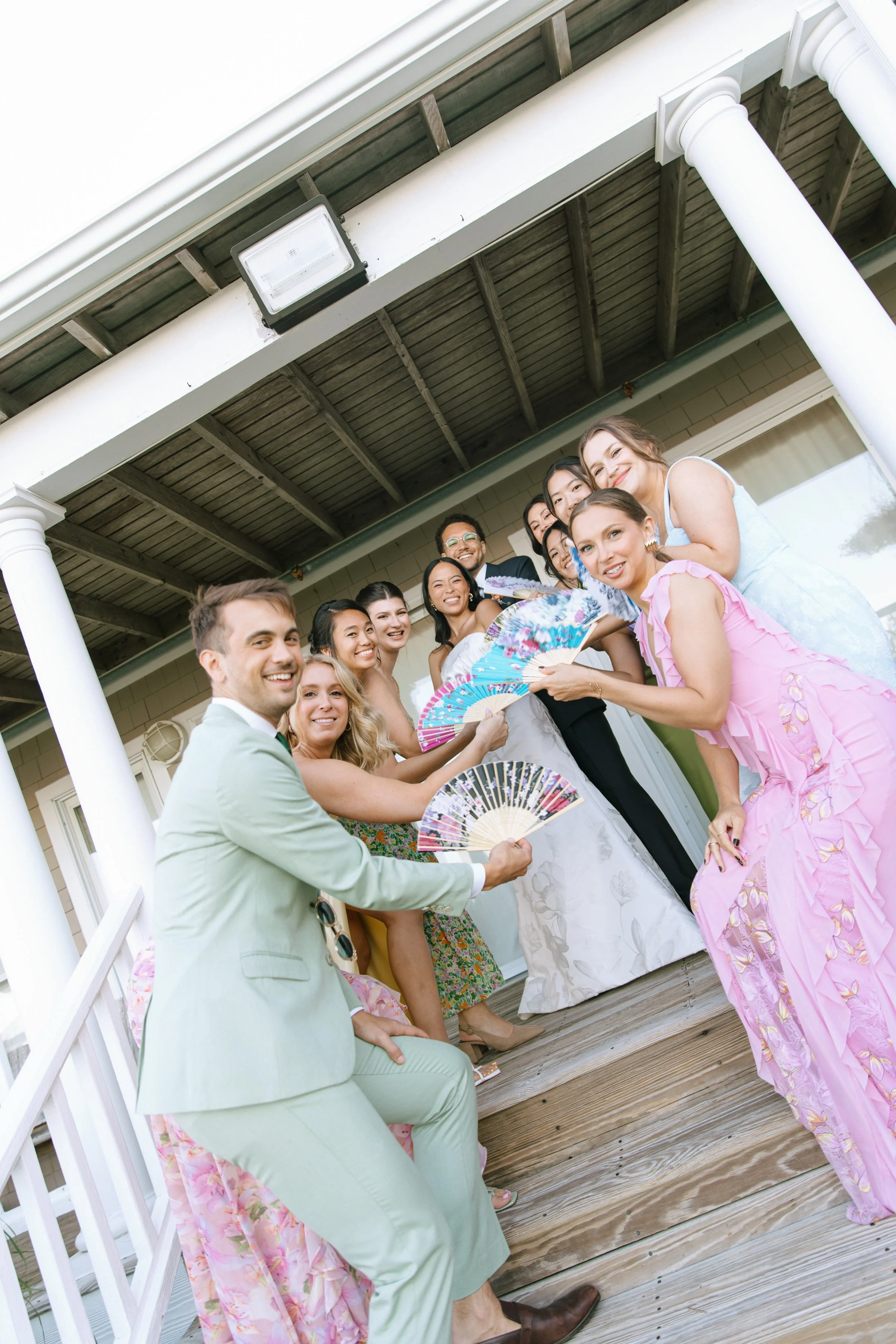Enjoying a celebration or gathering on a porch with friends, some women are holding decorative fans and all are smiling and dressed in colorful, festive attire.