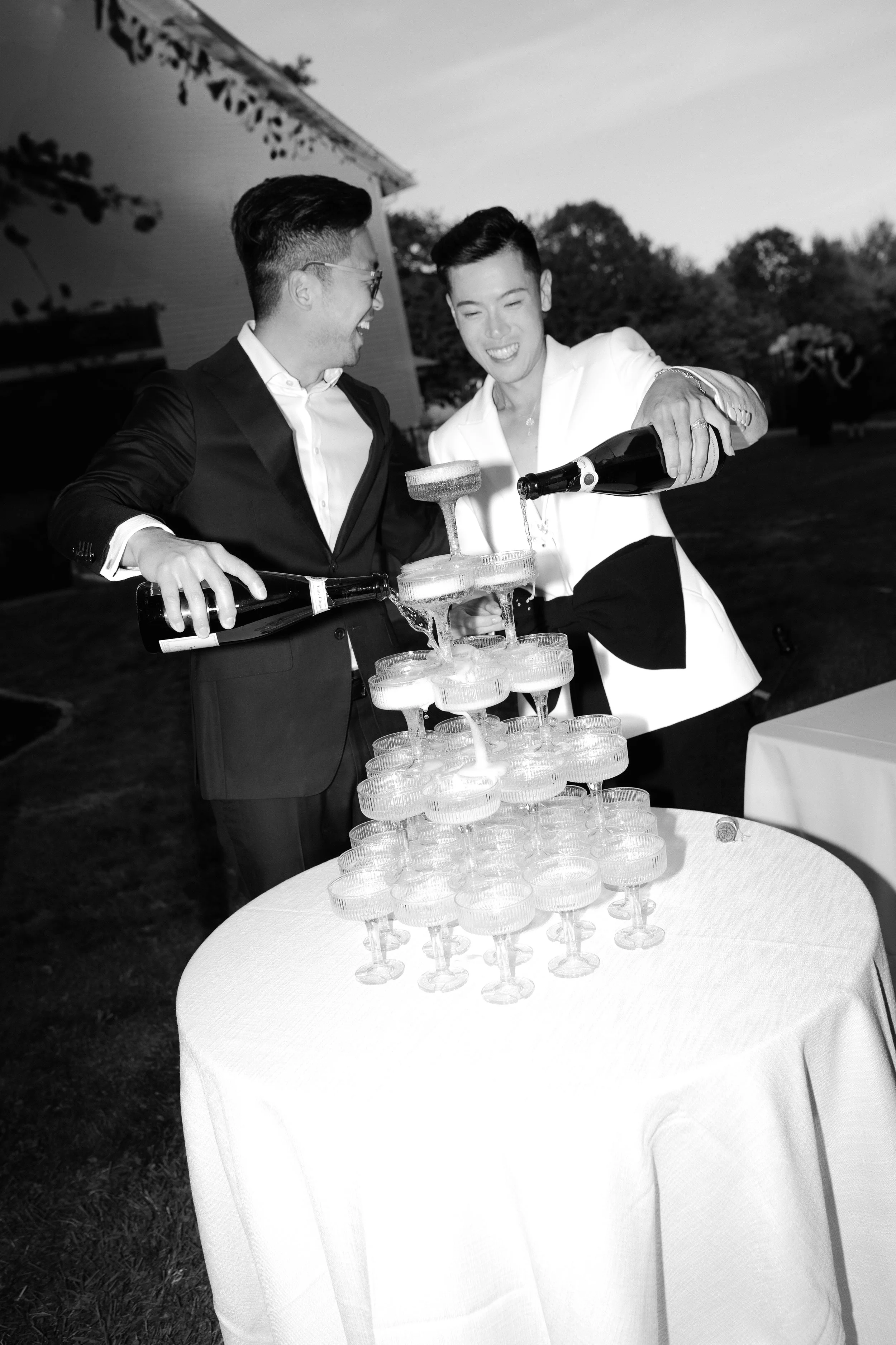 Two men in formal suits celebrating with a champagne tower at an outdoor event.