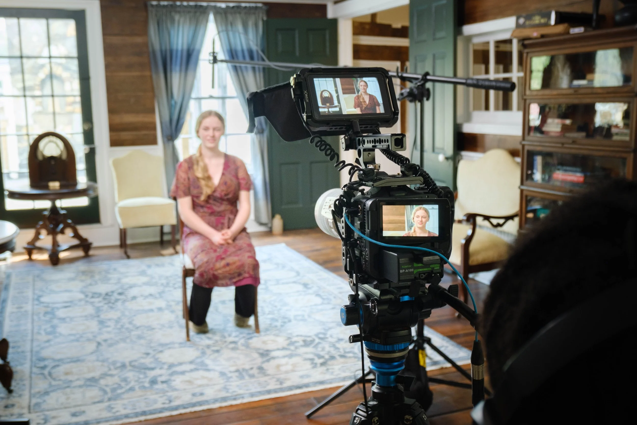 A person operating a professional video camera captures an interview scene of a woman sitting on a chair indoors with wooden walls, windows, and furniture in the background.