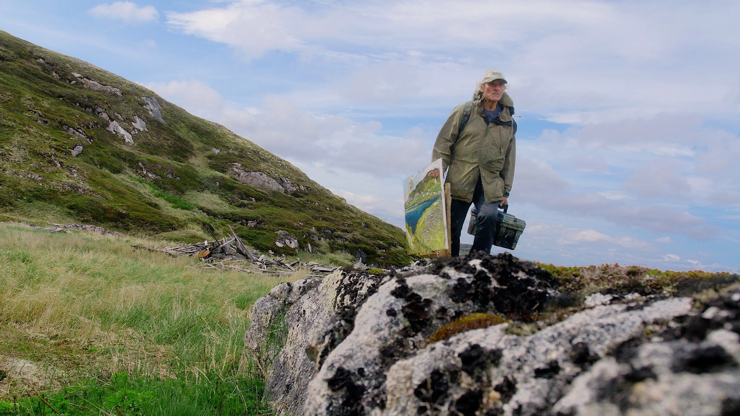 An artist carrying a painting and a box walks on a grassy field near a hillside.