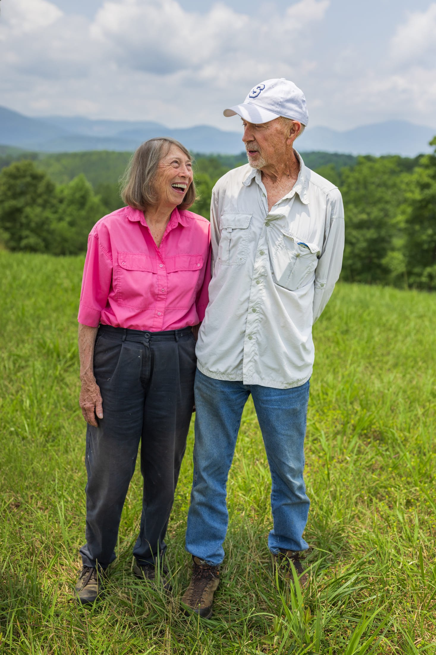An elderly couple smiling and talking while standing in a lush green field with mountains and cloudy sky in the background.