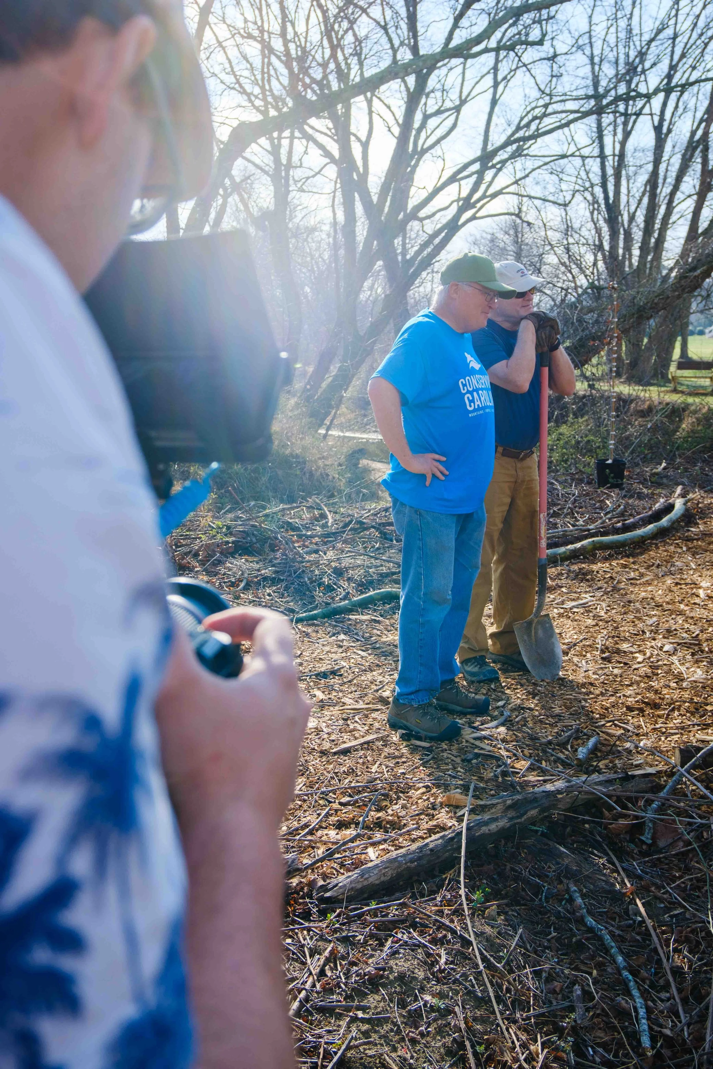 Three men in a wooded outdoor area with fallen branches, one holding a shovel, appear to be participating in a tree cleanup or community service event on a sunny day.