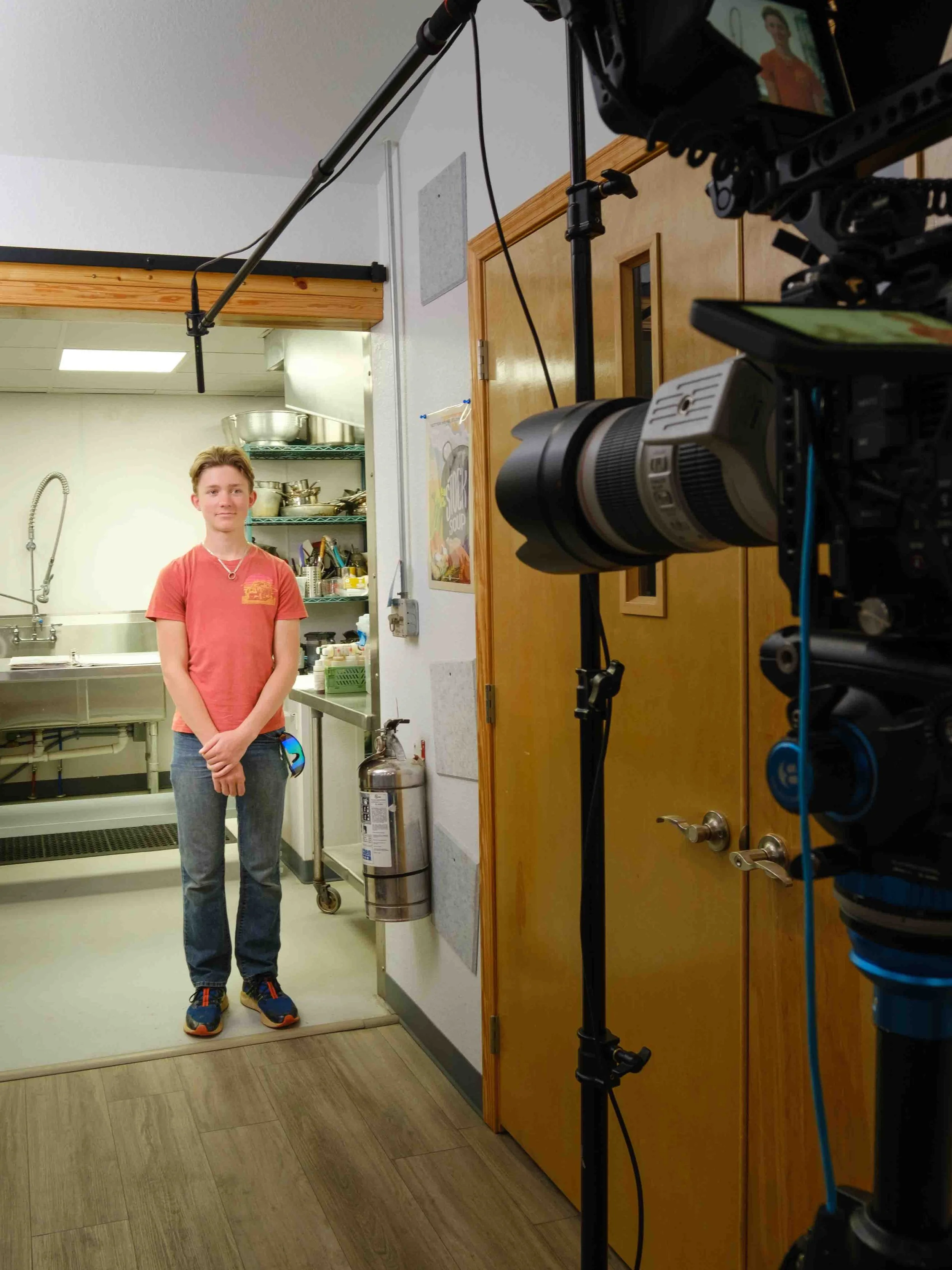 A young man stands in a kitchen behind a filming set with professional camera equipment, including a camera on a tripod, capturing his portrait. The kitchen has a stainless steel sink, shelves with cookware, and a fire extinguisher on the wall.
