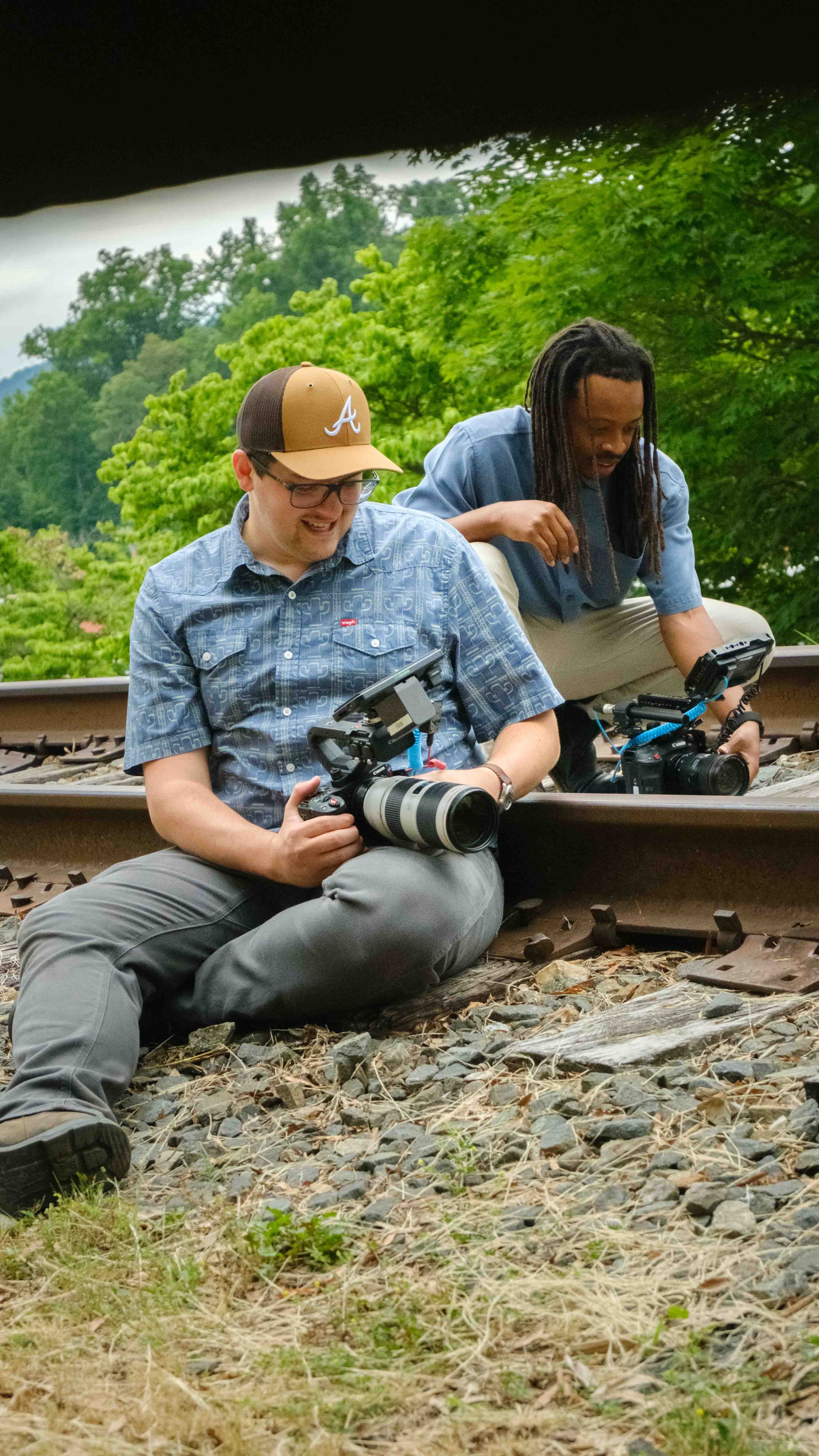 Two men sitting on a train track with cameras, surrounded by green trees and rocks.
