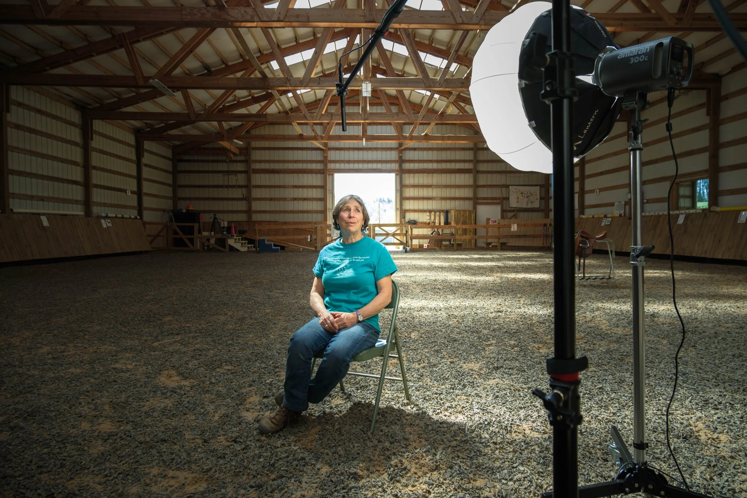 A woman sitting on a chair in a large indoor riding arena, with a photography light setup in front of her, and a wooden-fenced horse riding area in the background.