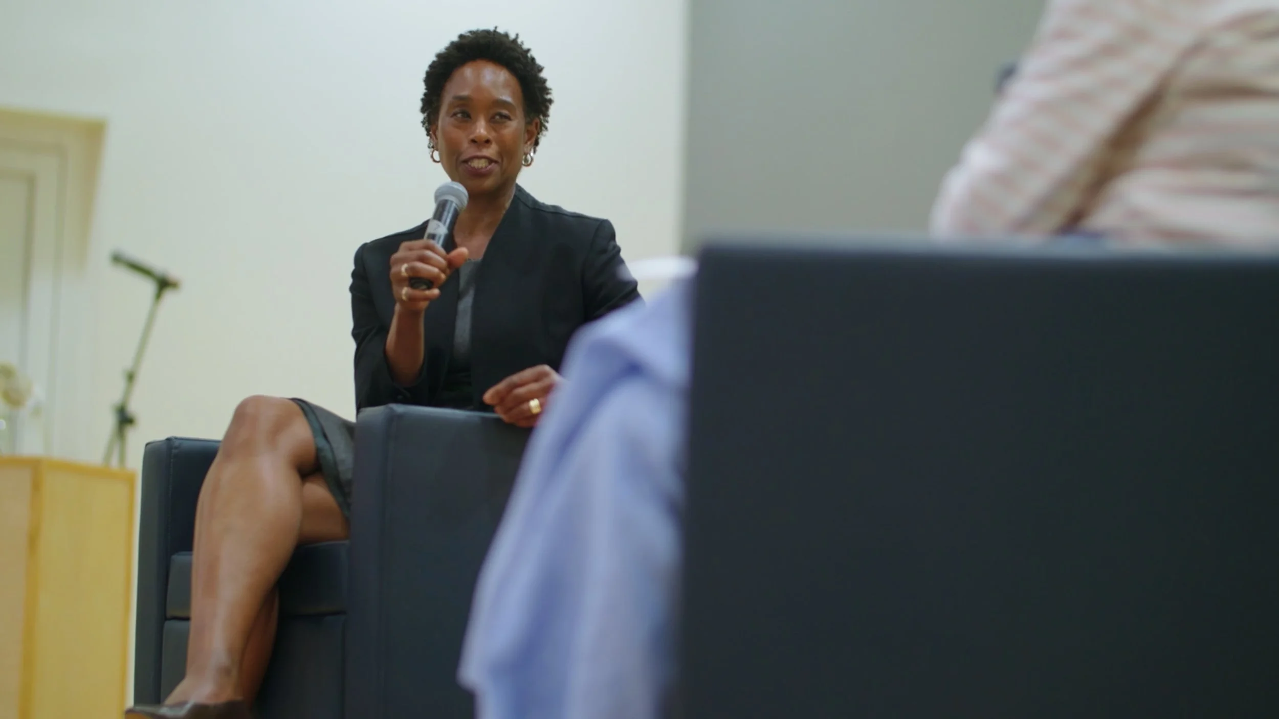 A woman holding a microphone and speaking during an interview or presentation, sitting on a chair in a formal setting.