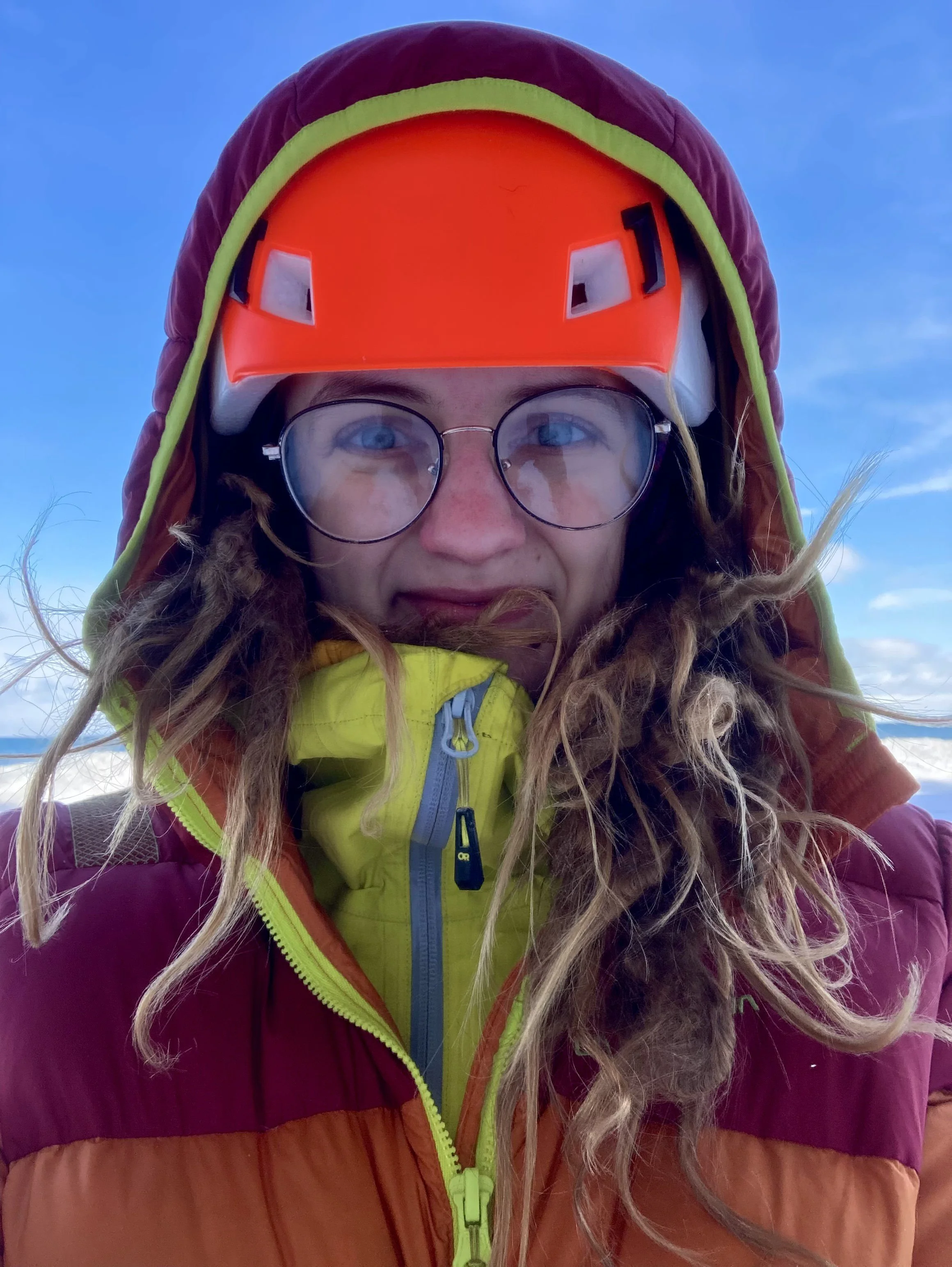 A woman wearing a bright orange helmet and a purple jacket with green accents, outdoors with blue sky, taking a close-up selfie.