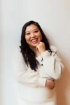 A smiling woman with long dark hair, wearing a cream cozy sweater, standing against a plain white wall.