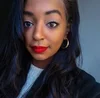 A young woman with dark wavy hair, red lipstick, and gold hoop earrings takes a close-up selfie against a neutral background.